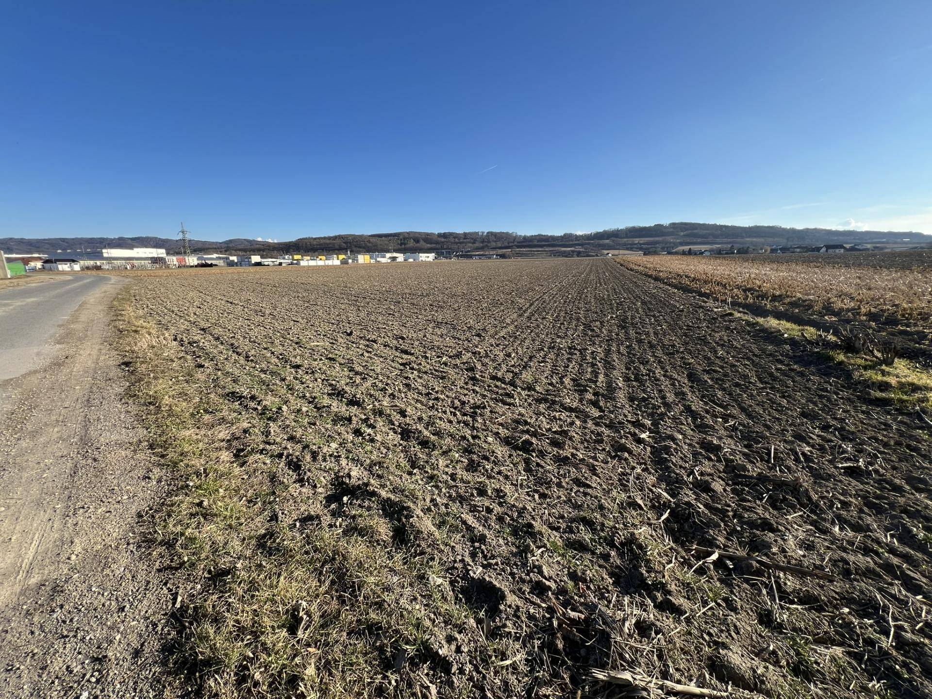 Weites Ackerland unter strahlendem Sonnenschein, mit einer Straße am Rand und Industriegebäuden in der Ferne.