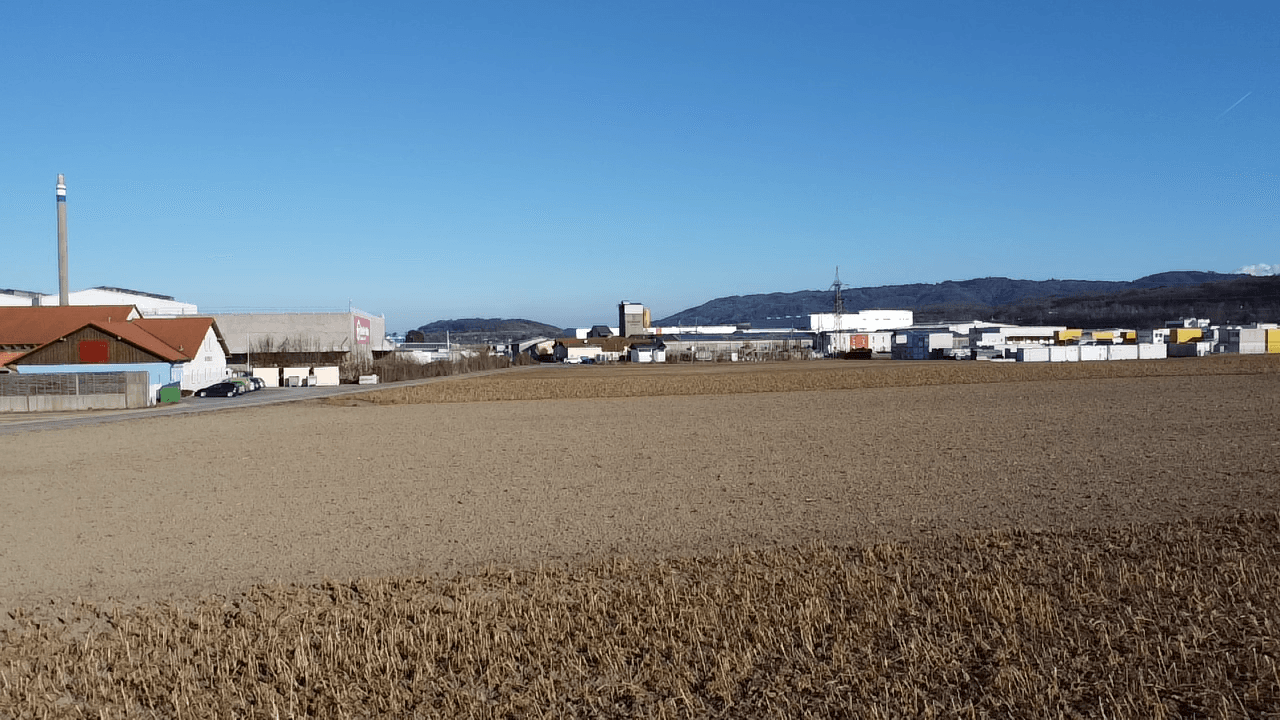 Weites Feld mit Blick auf Industriegebäude und Hügel in der Ferne unter blauem Himmel.