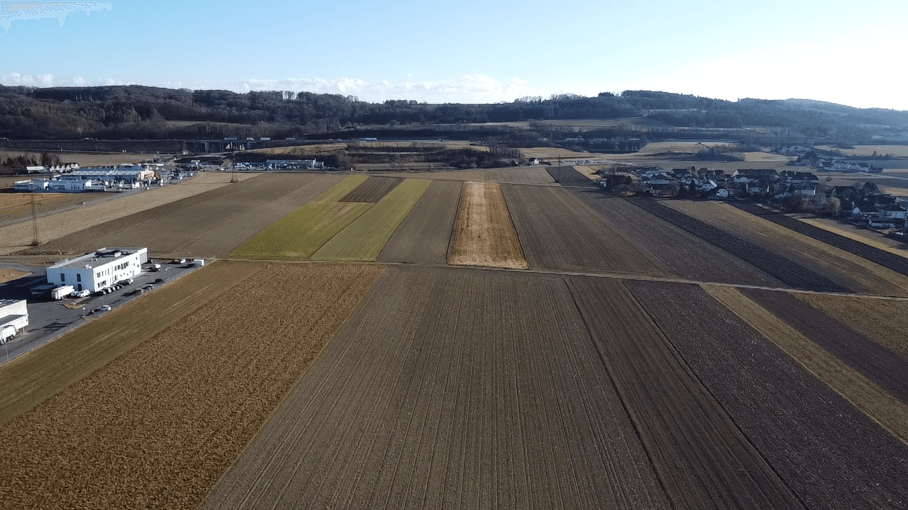 Weitläufige Ackerflächen erstrecken sich bis zum Horizont, mit Industriegebäuden und Hügeln in der Ferne.