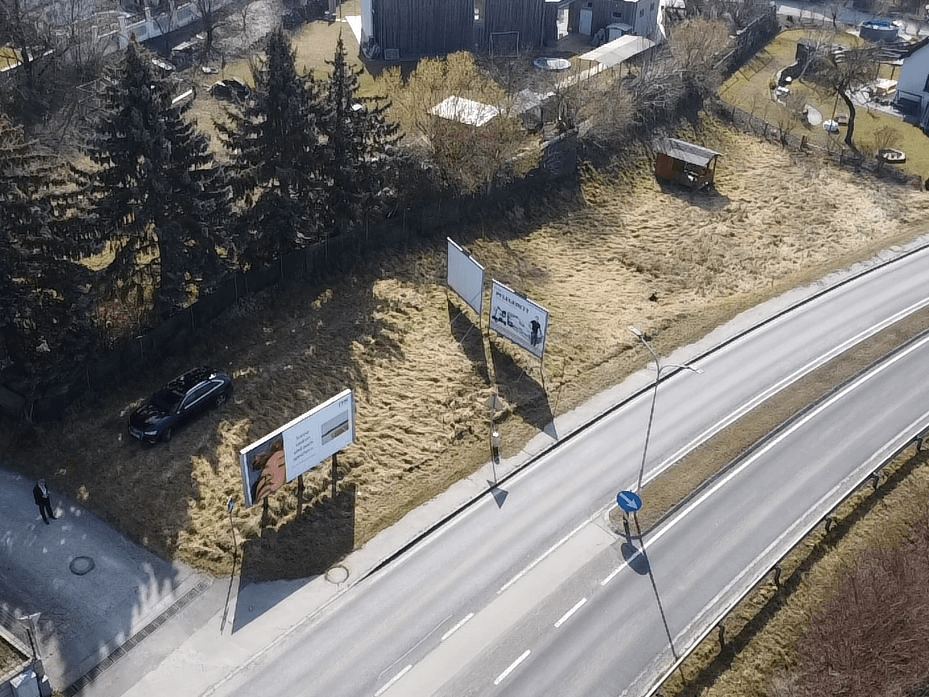 Übersicht aus der Vogelperspektive eines unbebauten Grundstücks an einer Straße, mit Werbetafeln und einem Fahrzeug.