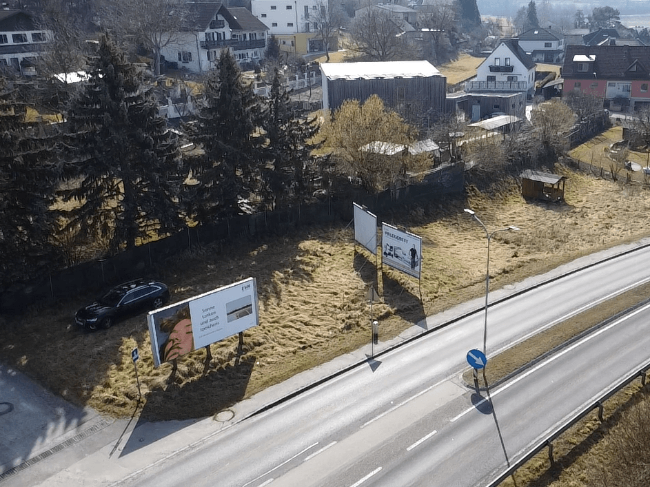 Luftaufnahme eines unbebauten Grundstücks neben einer Straße, mit einem geparkten Auto und Werbetafeln.