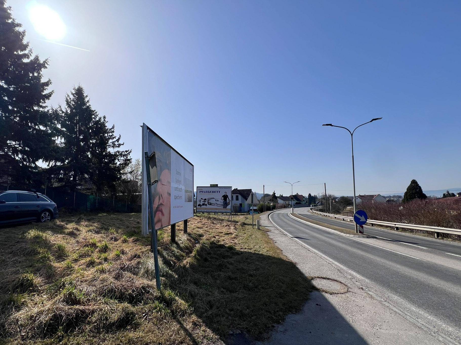 Blick entlang der Straße auf das Grundstück mit einer Werbetafel und der ländlichen Umgebung an einem sonnigen Tag.