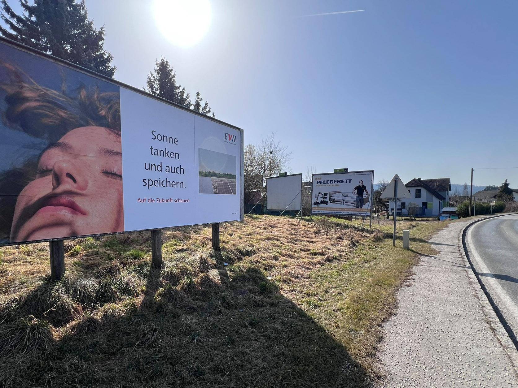 Ansicht des Grundstücks mit zwei Werbetafeln, einem geparkten Auto und der Straße im Hintergrund bei Sonnenschein.