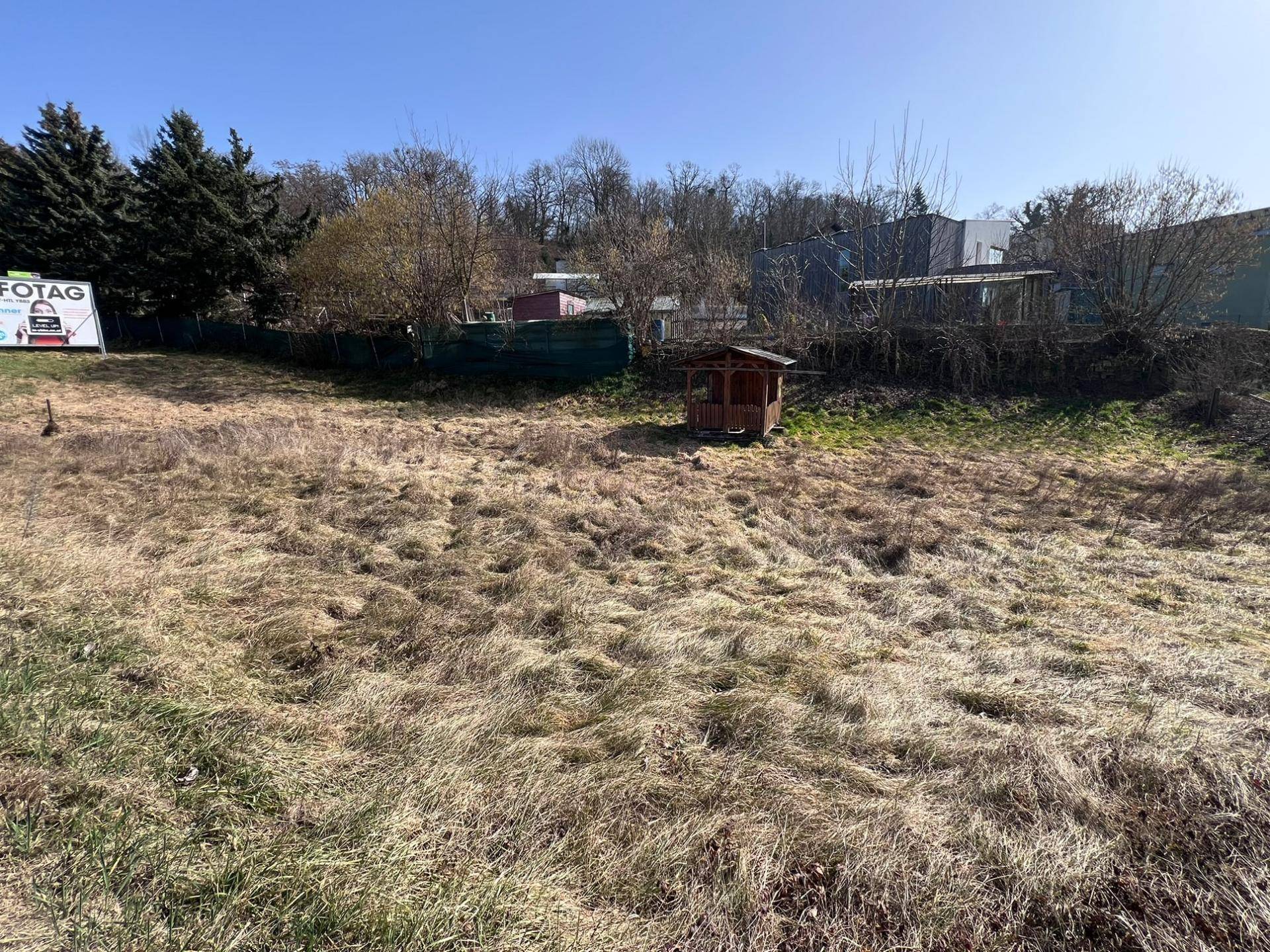 Blick über das grasbewachsene Grundstück mit einem Holzpavillon und Bäumen im Hintergrund an einem sonnigen Tag.