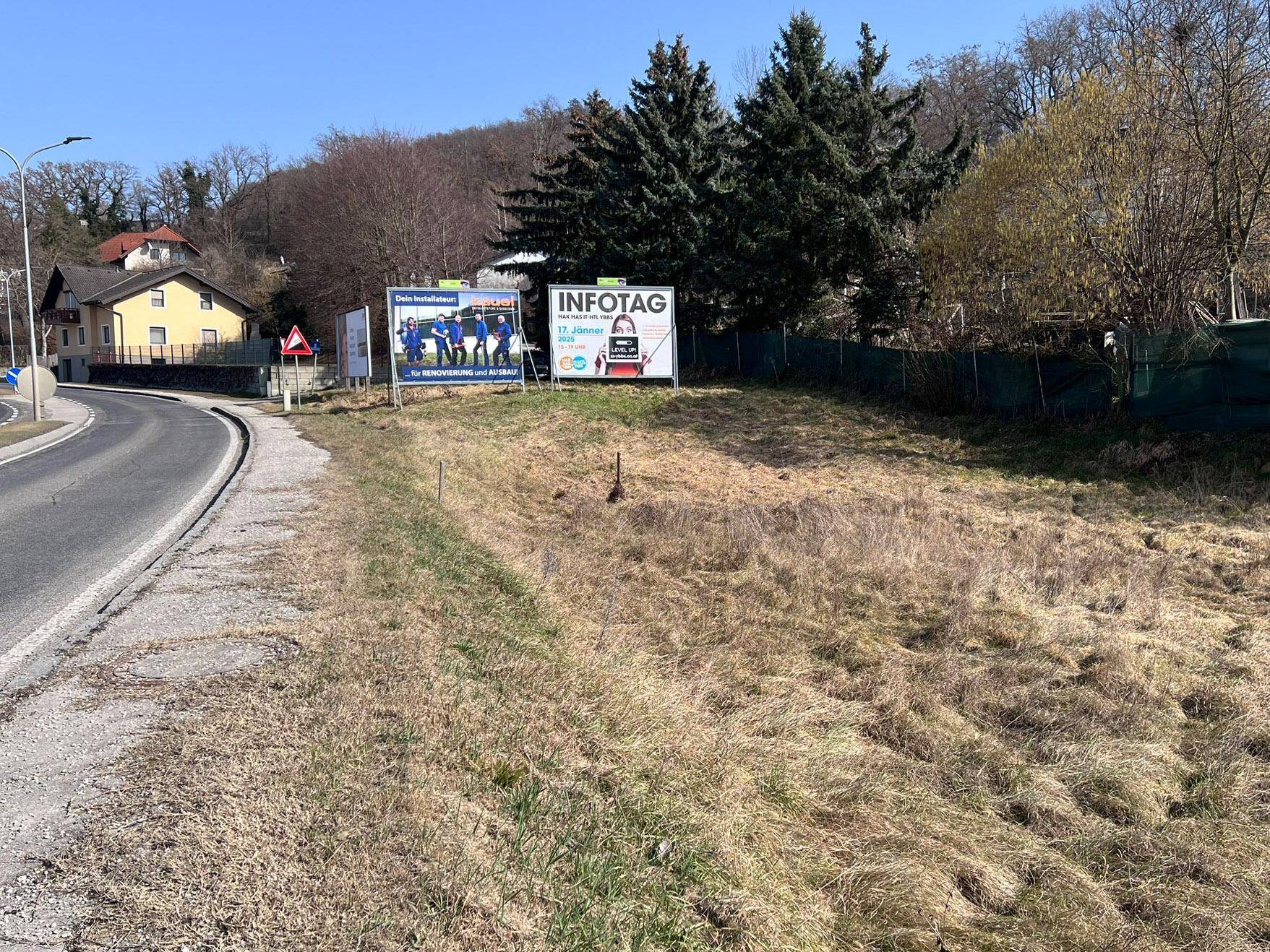 Blick über das Grundstück mit zwei Werbetafeln, der Straße und Häusern in der Ferne an einem sonnigen Tag.