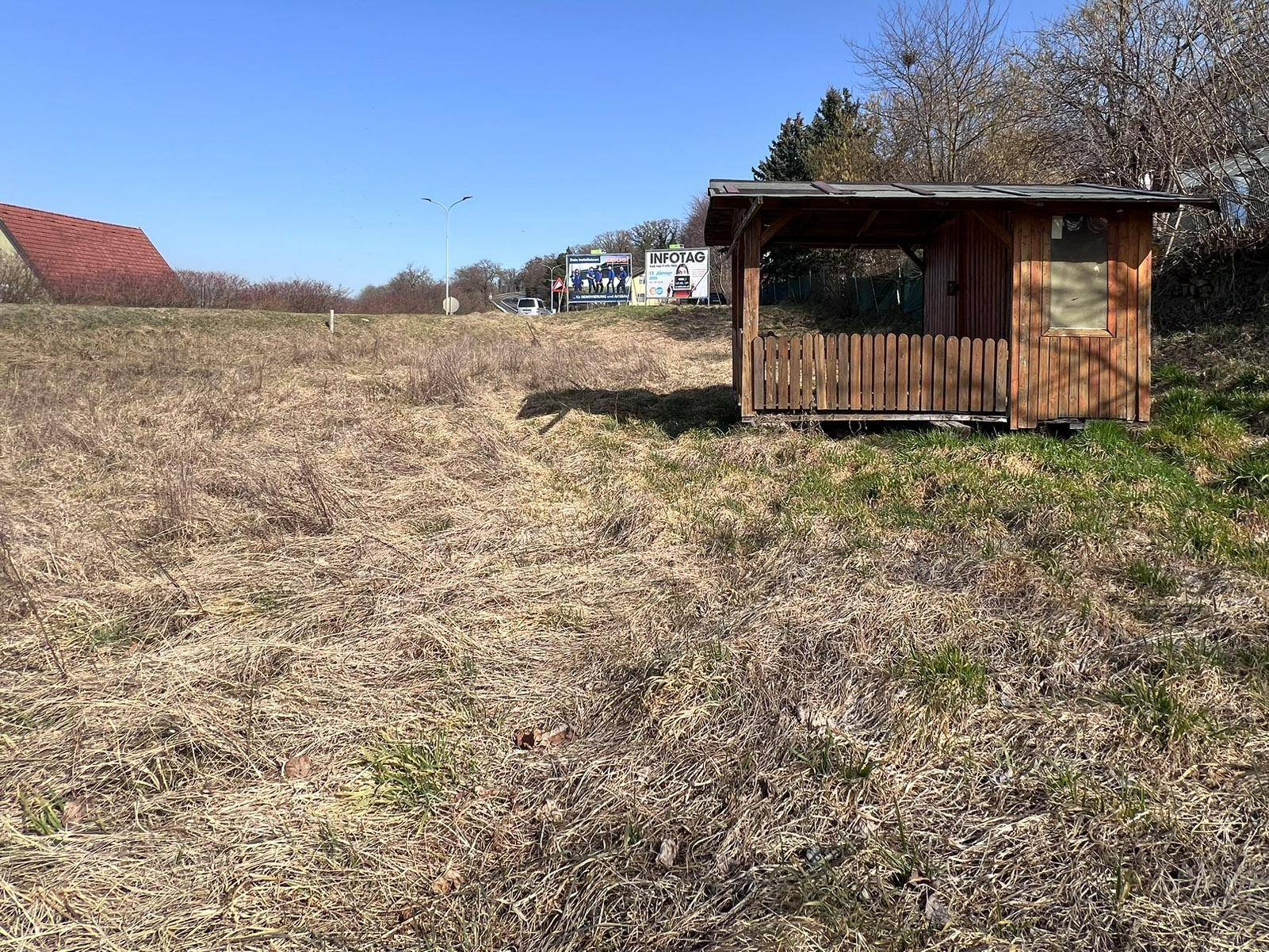 Holzpavillon auf dem Grundstück mit einer Werbetafel im Hintergrund und der ländlichen Umgebung bei Sonnenschein.