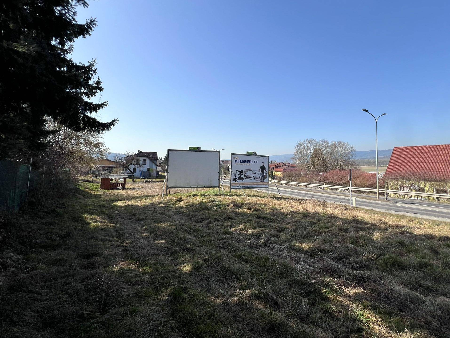 Weitwinkelaufnahme des Grundstücks mit zwei Werbetafeln, der Straße und der ländlichen Hügellandschaft im Hintergrund.