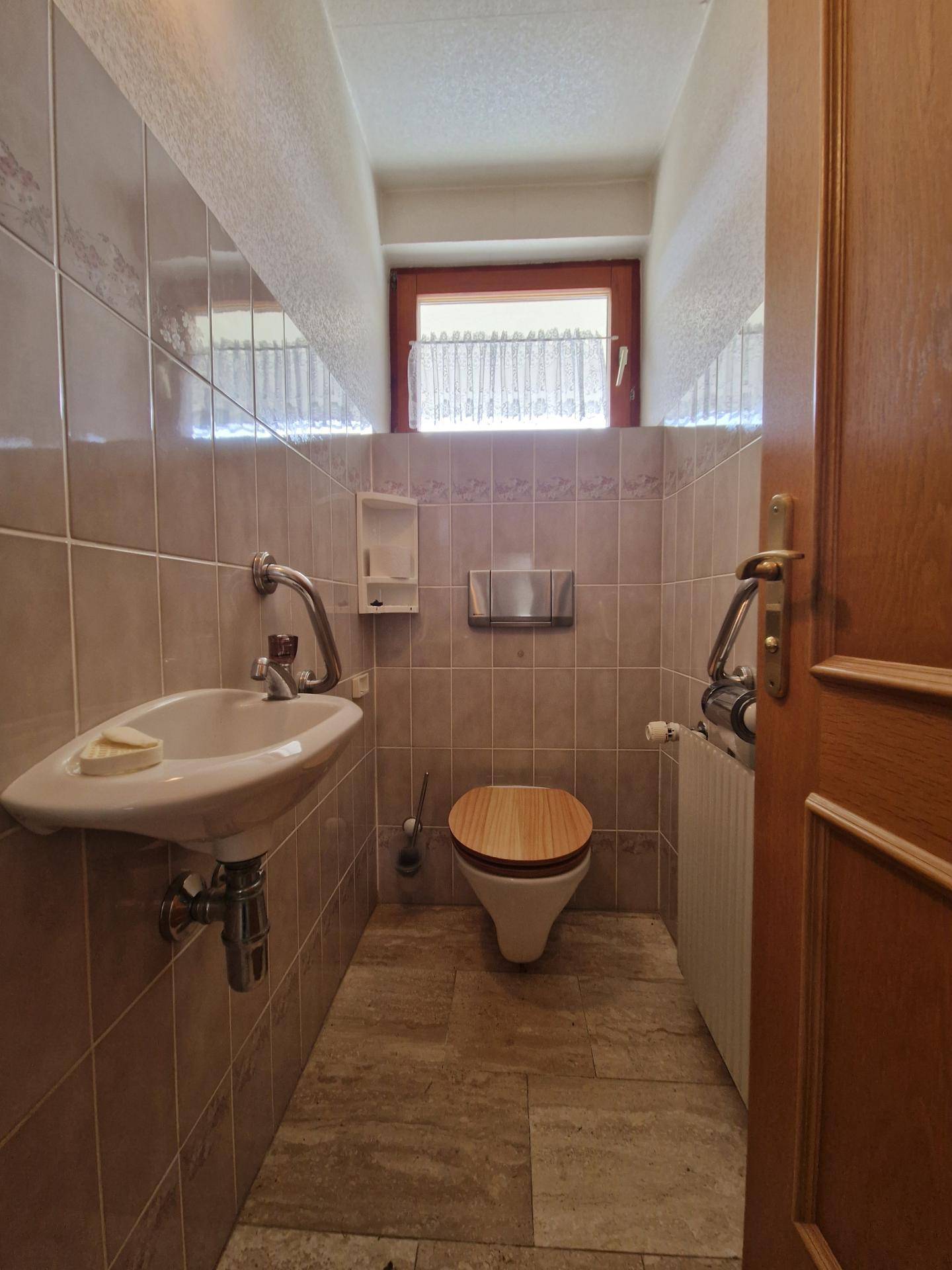 Separate Toilette mit Fenster, Waschbecken und Fliesen an Wänden und Boden.