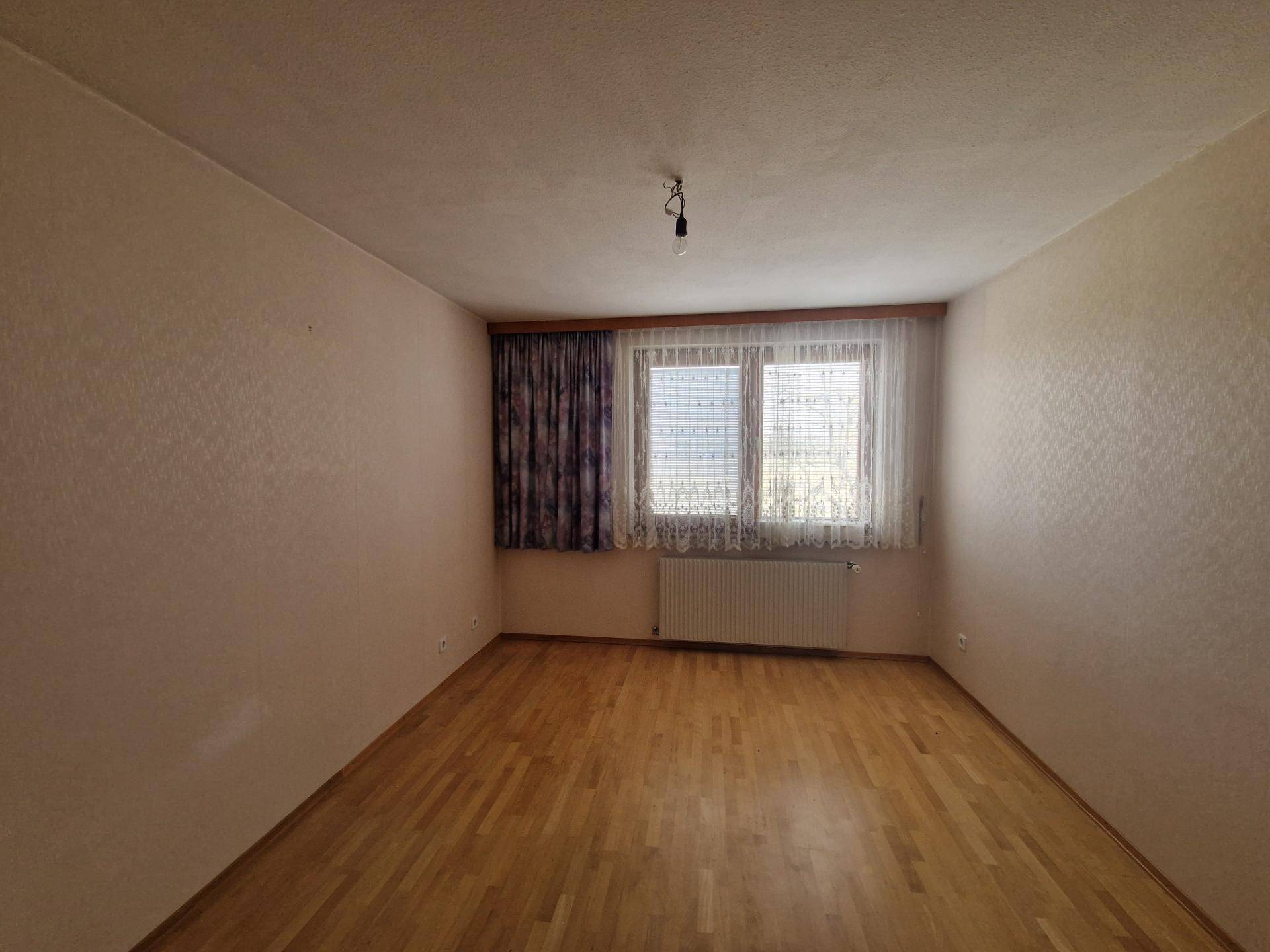 Helles Schlafzimmer mit Parkettboden und Fenster mit gemusterten Vorhängen.