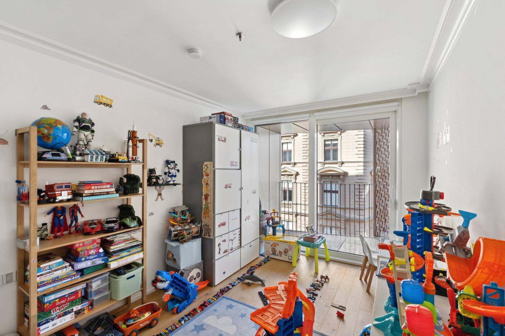 Helles Kinderzimmer mit vielen Spielsachen, Regalen und Zugang zu einem französischen Balkon.
