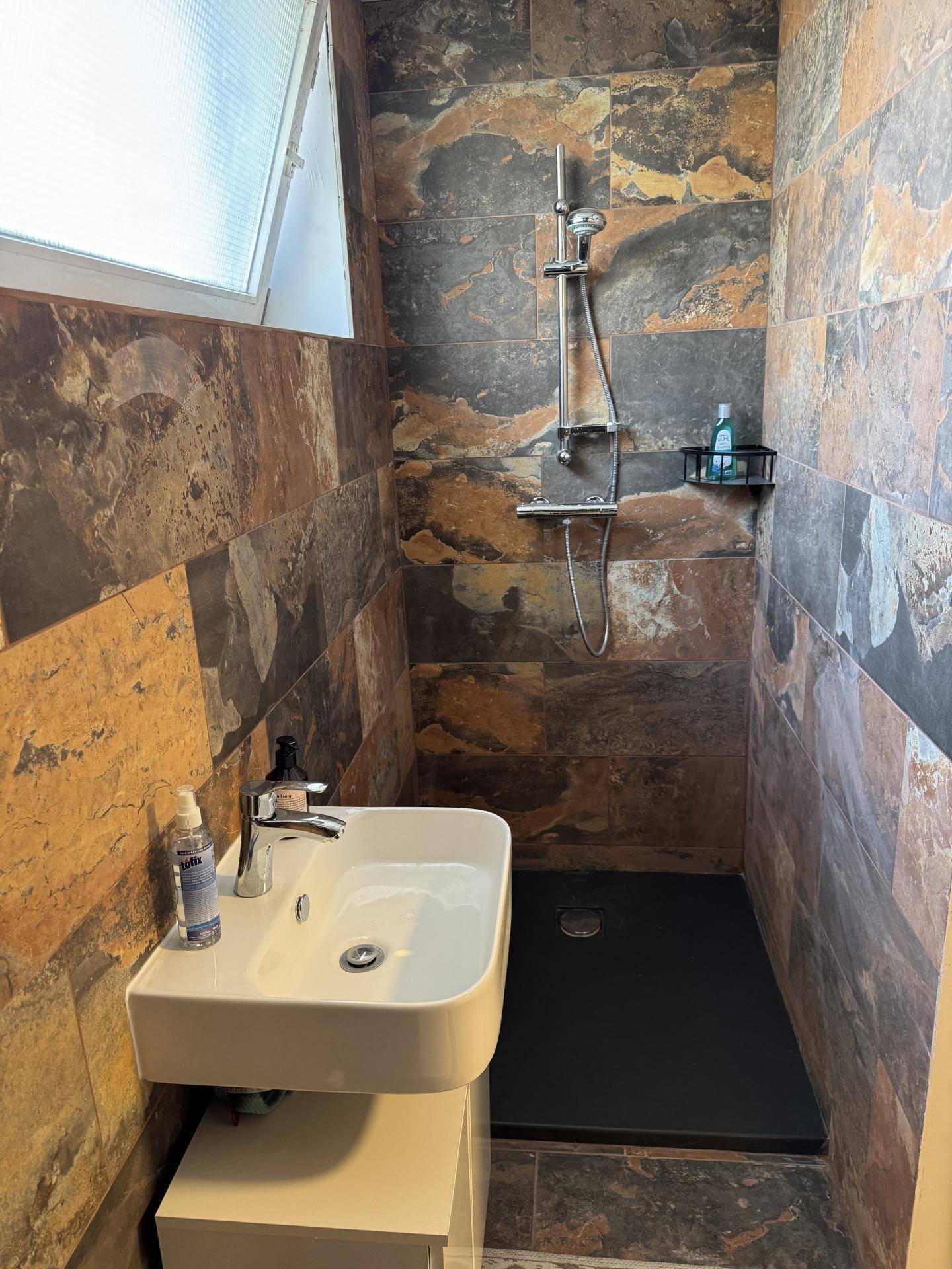 Modernes Badezimmer mit begehbarer Dusche, kleinem Waschbecken und Natursteinfliesen.