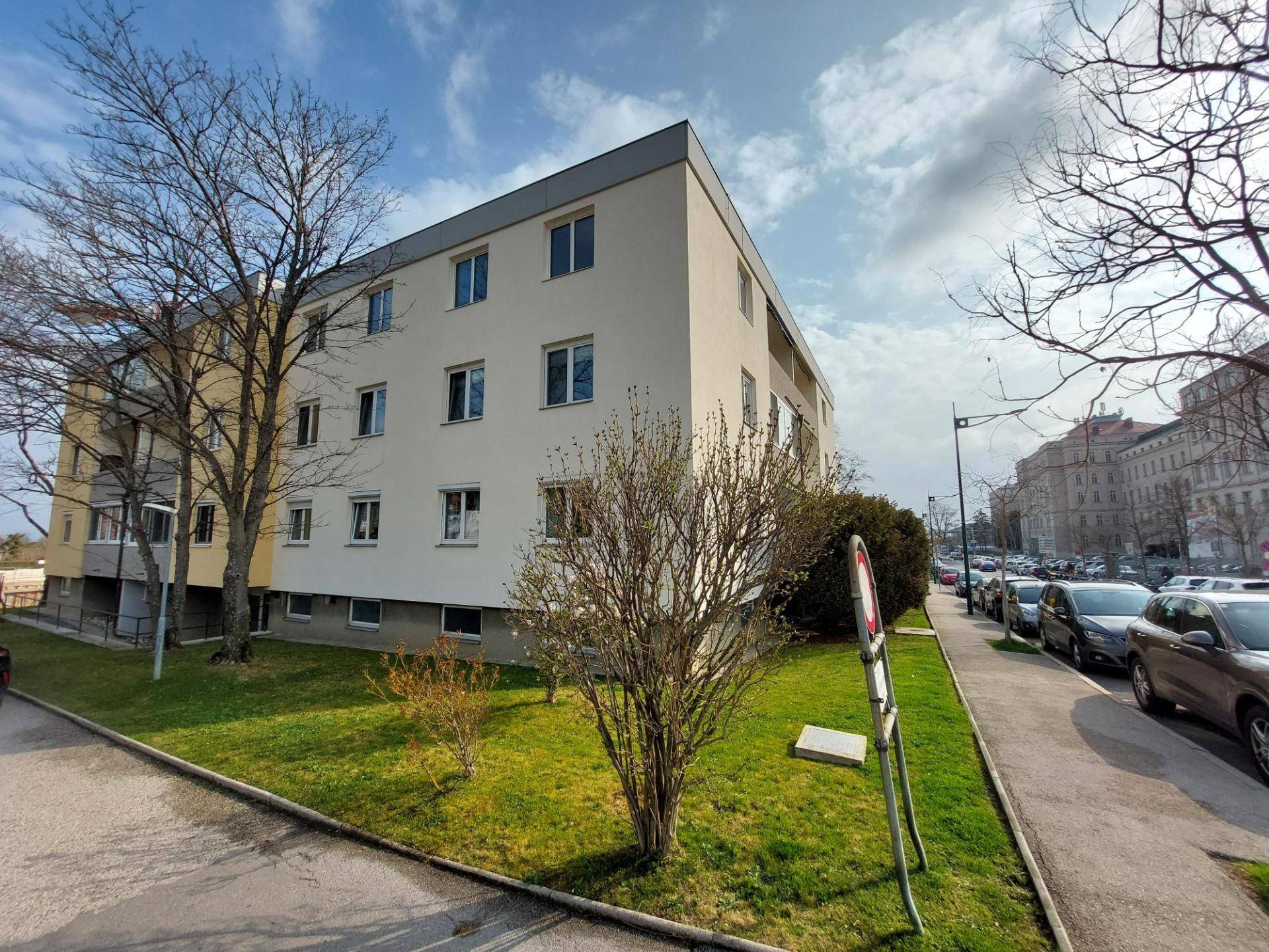 Ansicht eines Mehrfamilienhauses mit heller Fassade, Grünflächen und Bäumen an einem sonnigen Tag.