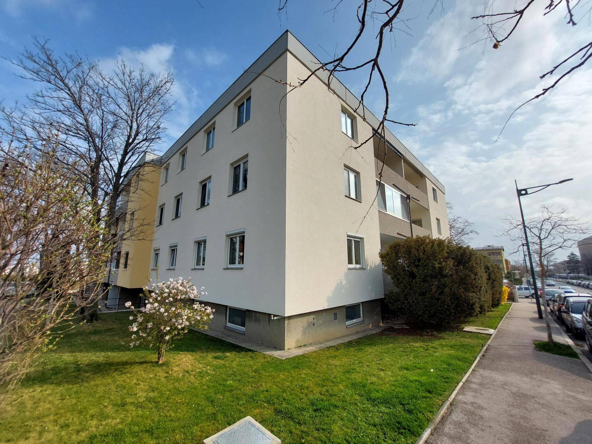 Mehrfamilienhaus mit heller Fassade, gepflegten Grünflächen und blühendem Baum.