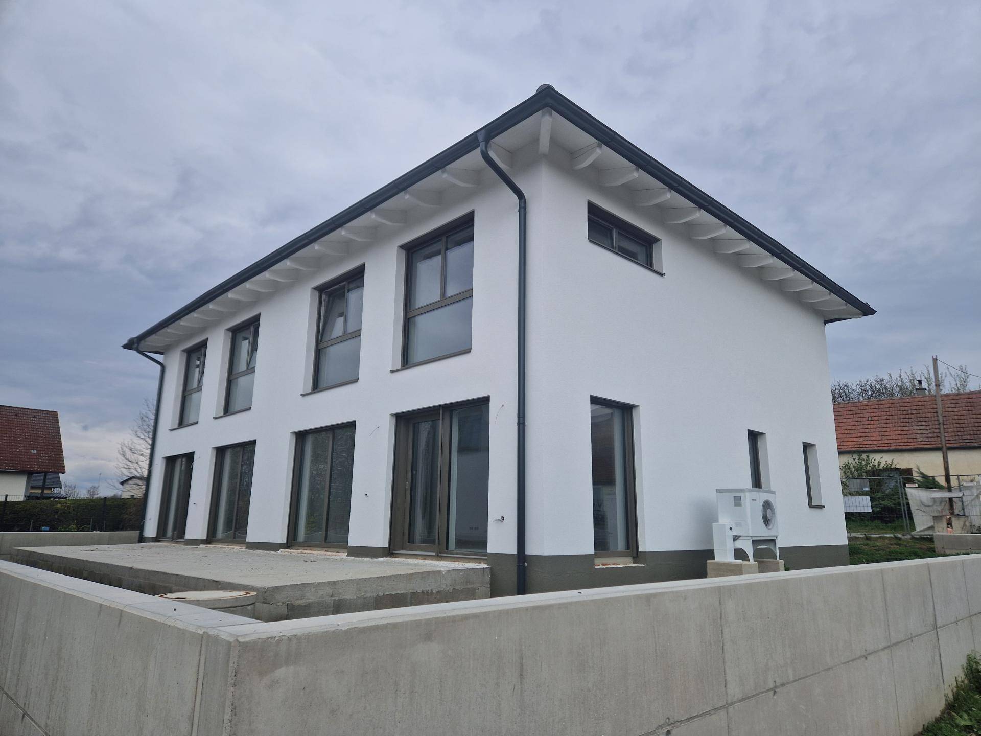 Moderne Doppelhaushälfte im Bau mit großen Fensterfronten, weißer Putzfassade und Wärmepumpe.