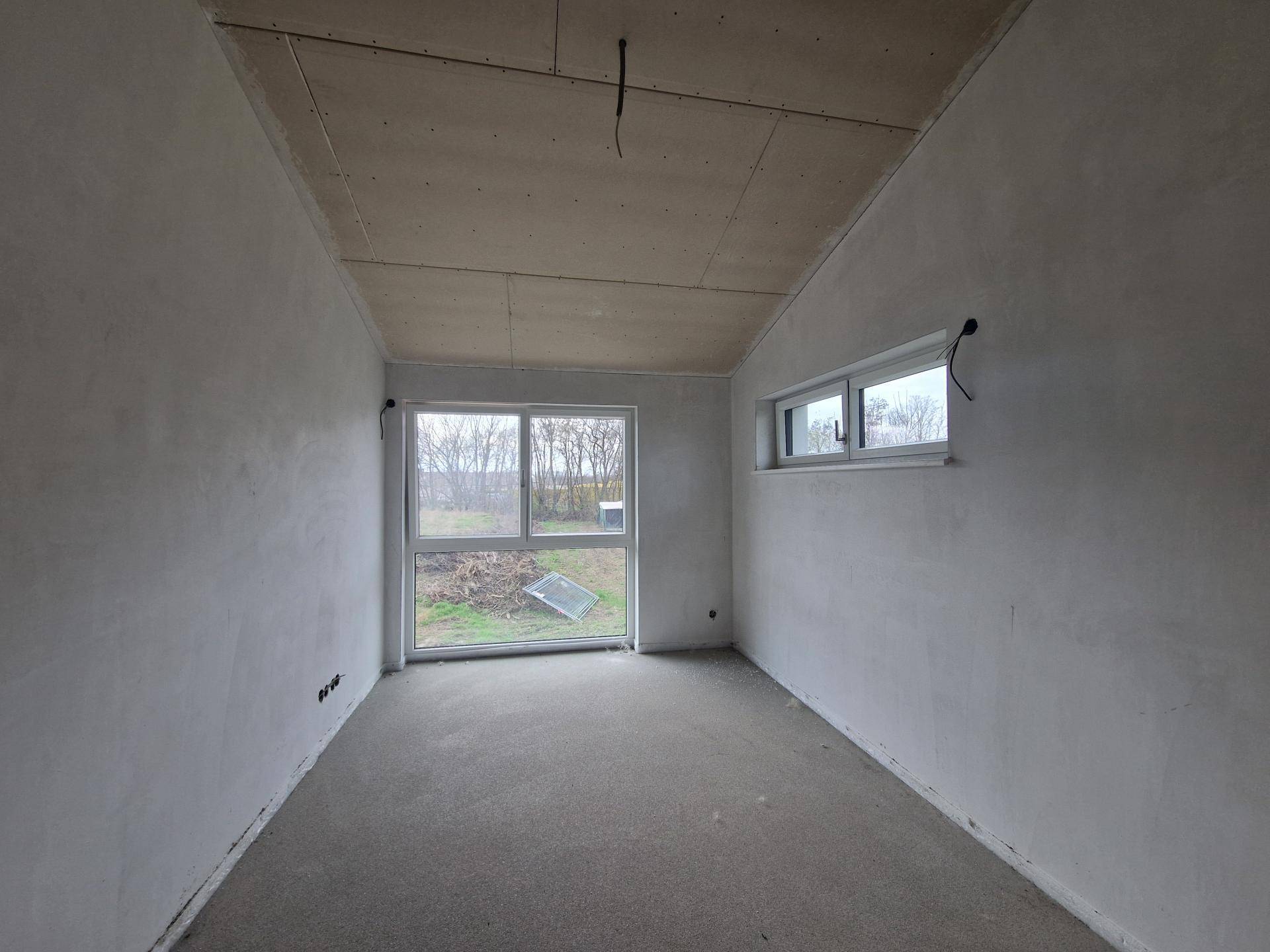 Unfertiges Zimmer mit zwei Fenstern, darunter eine große Glasfront, die den Blick ins Grüne freigibt.