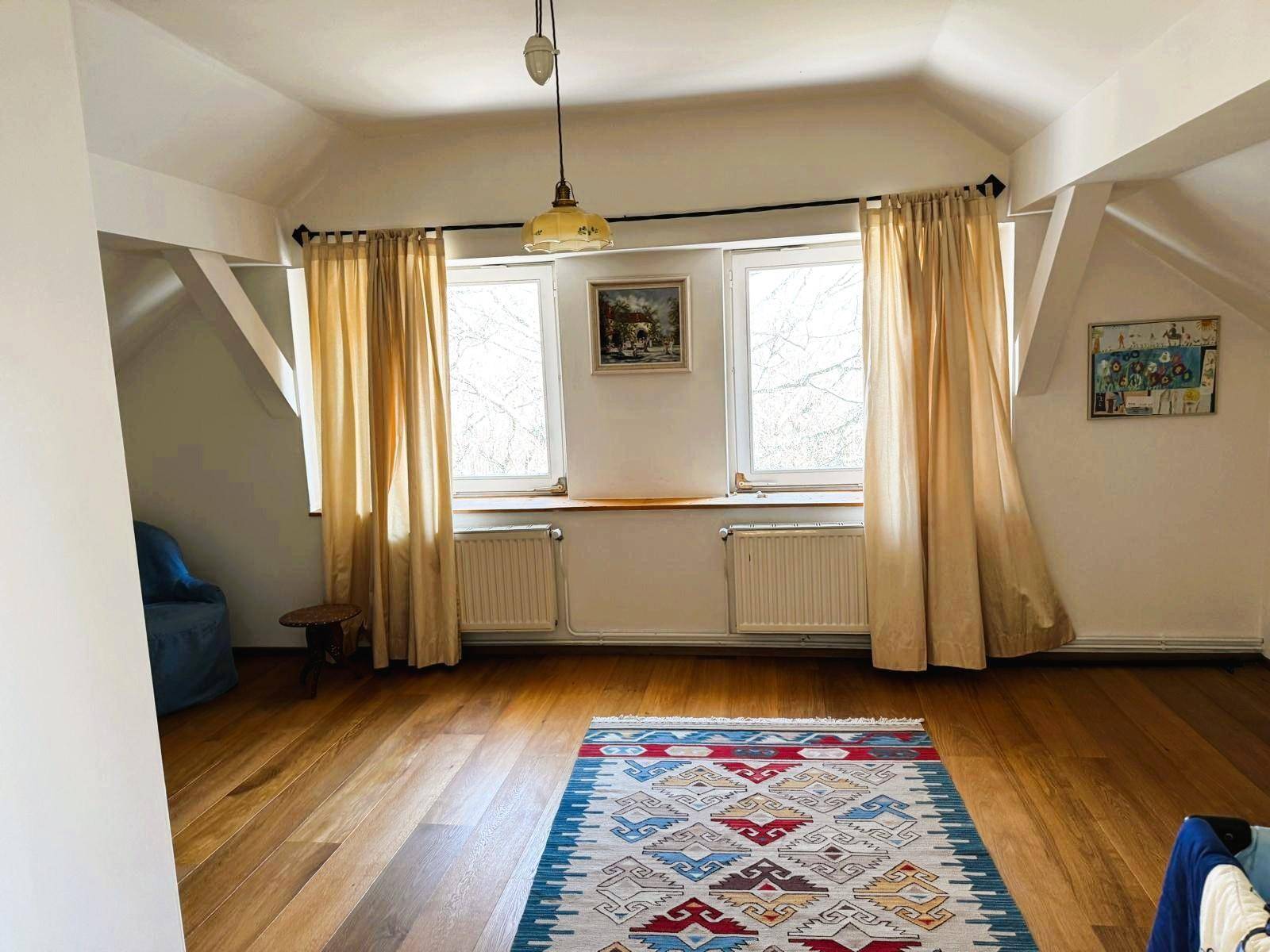Geräumiges Dachgeschoss-Schlafzimmer mit Holzboden, zwei Fenstern und einem bunten Teppich.