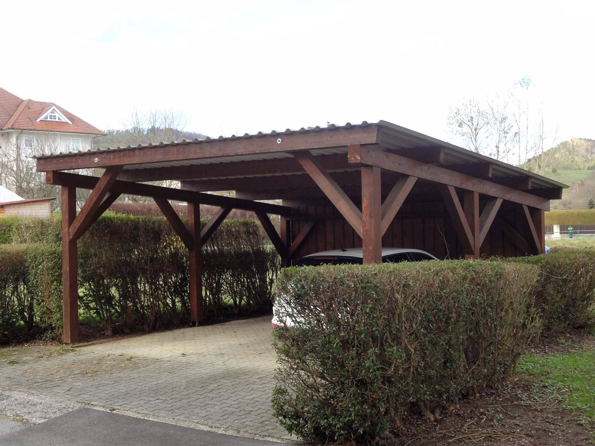 Holz-Carport für Fahrzeuge, umgeben von Hecken und Grünflächen.