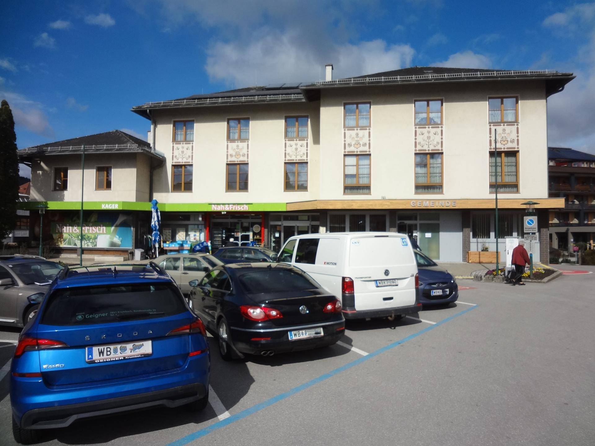 Mehrfamilienhaus mit Geschäften im Erdgeschoss und Parkplätzen davor.