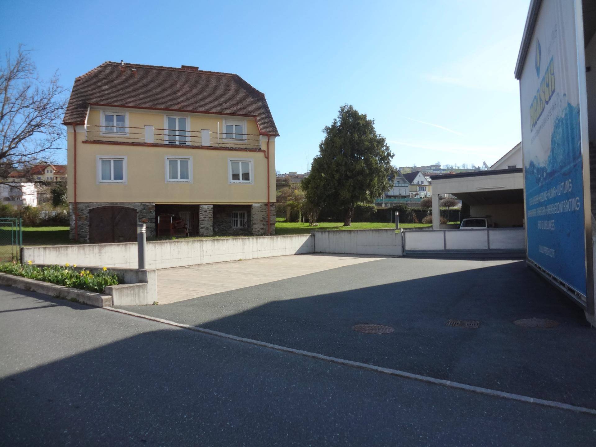 Außenansicht eines Gebäudes mit großem Parkplatz und angrenzendem Wohnhaus im Hintergrund.