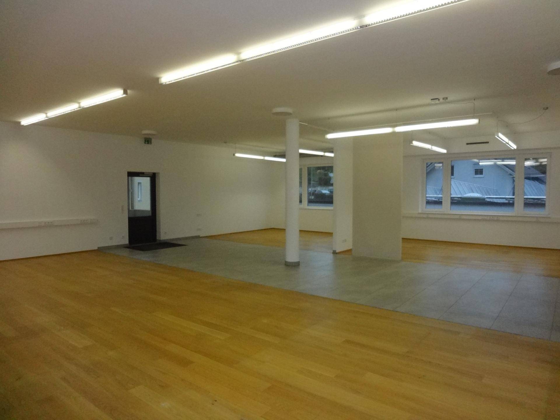 Weitläufiger Büroraum mit Holz- und Fliesenboden, ausgestattet mit Deckenleuchten und Fenstern.