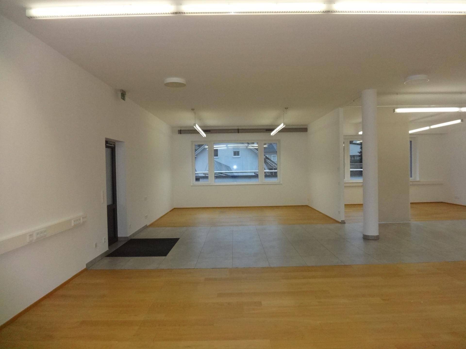 Geräumiger, leerer Büroraum mit großen Fenstern, die viel Tageslicht hereinlassen, und gemischtem Bodenbelag.