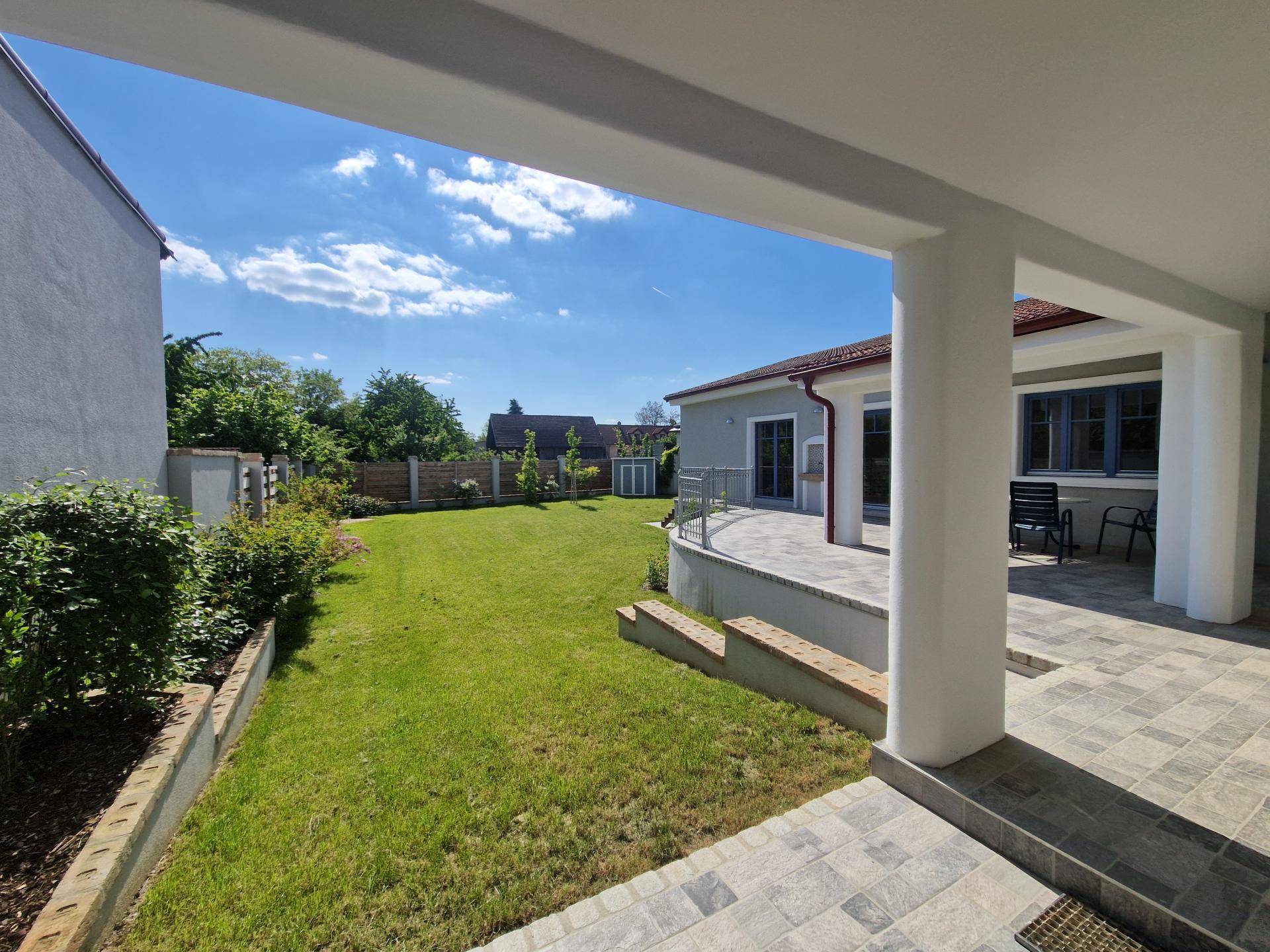 Überdachte Terrasse mit Fliesenboden und Blick auf den gepflegten Garten unter blauem Himmel.