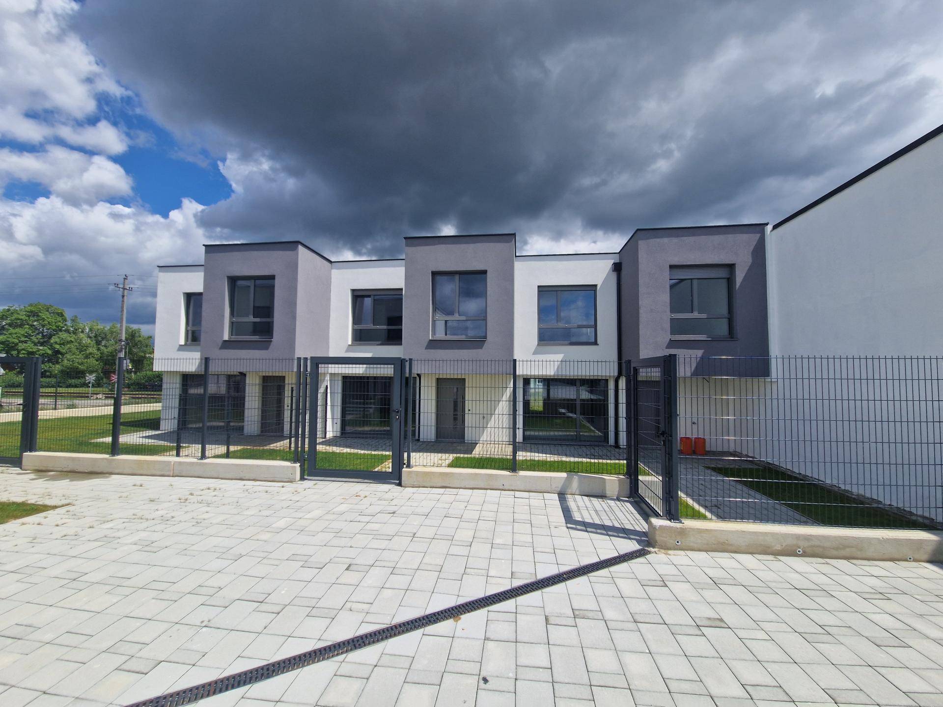Reihenhäuser mit moderner Architektur, kleinen Gärten und gepflastertem Zugangsbereich unter dunklen Wolken.
