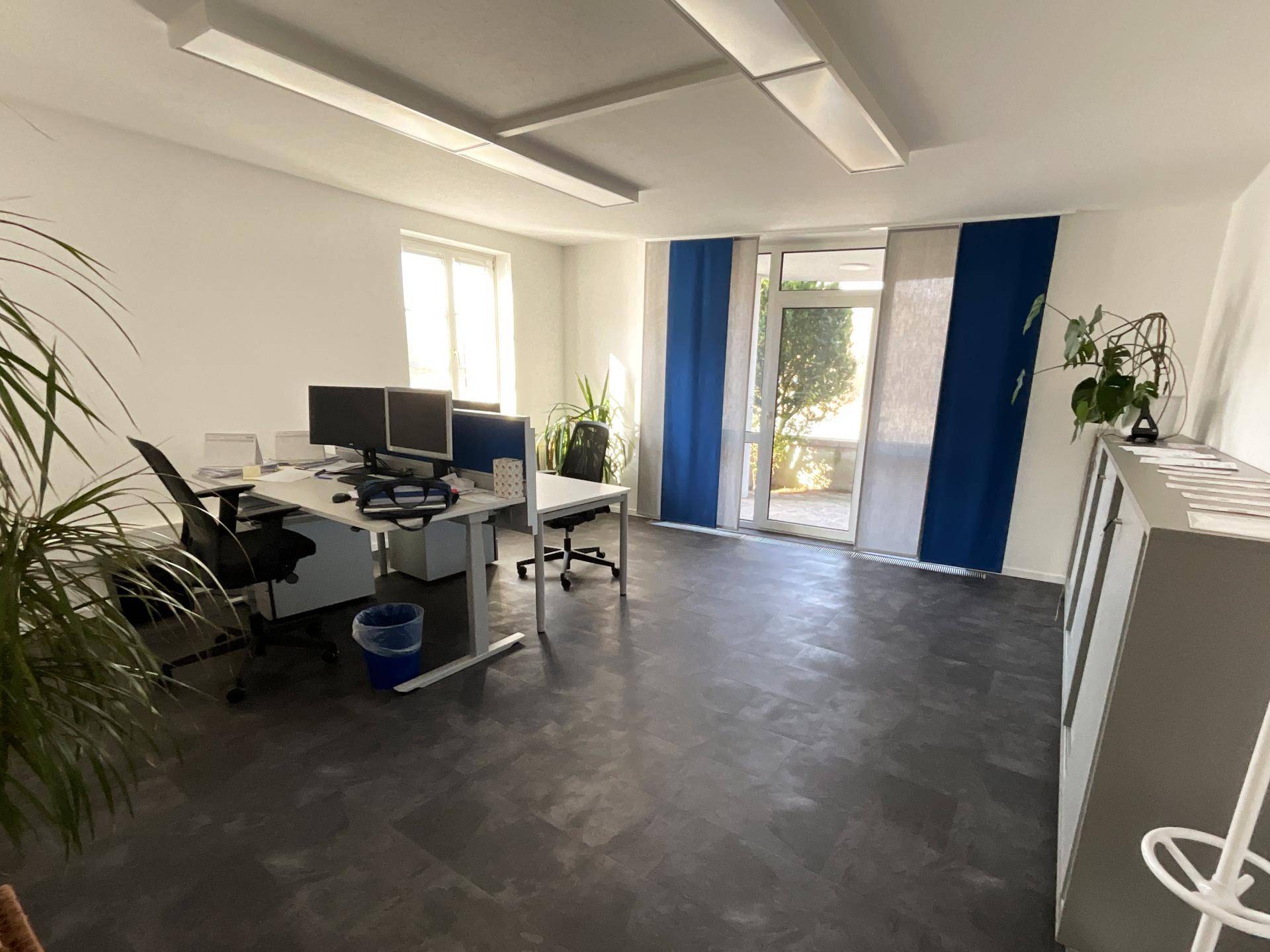 Geräumiges Büro mit mehreren Arbeitsplätzen, dunklem Boden und einer Glastür, die zu einem kleinen Außenbereich führt.