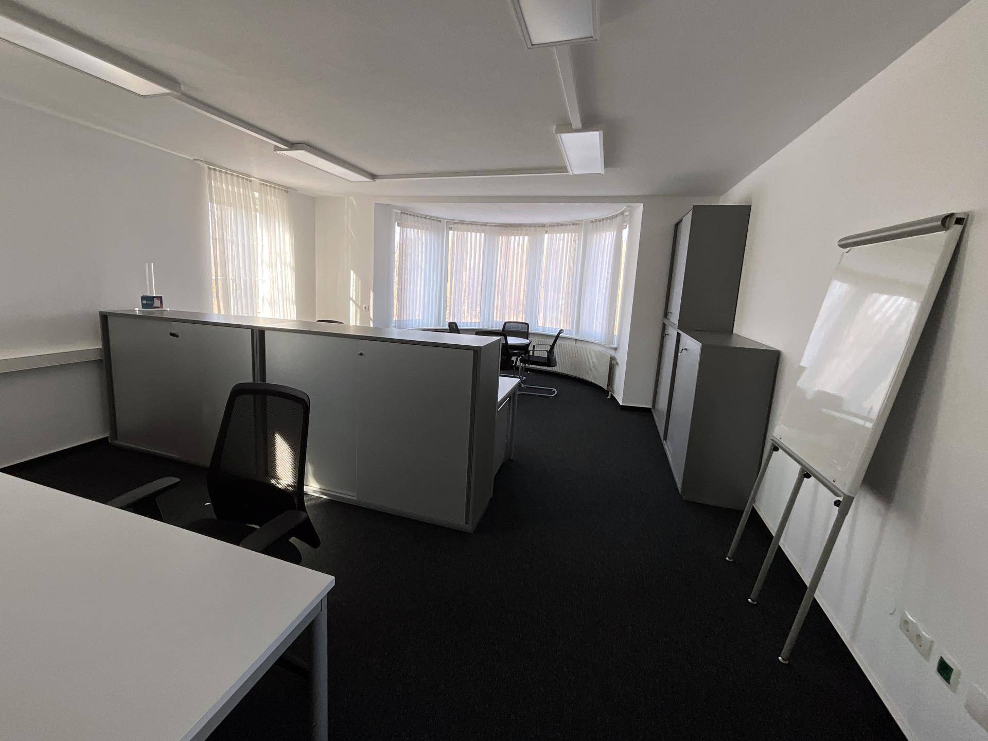 Großes Büro mit Teppichboden, mehreren Schreibtischen und einem Erkerfenster mit Vorhängen.