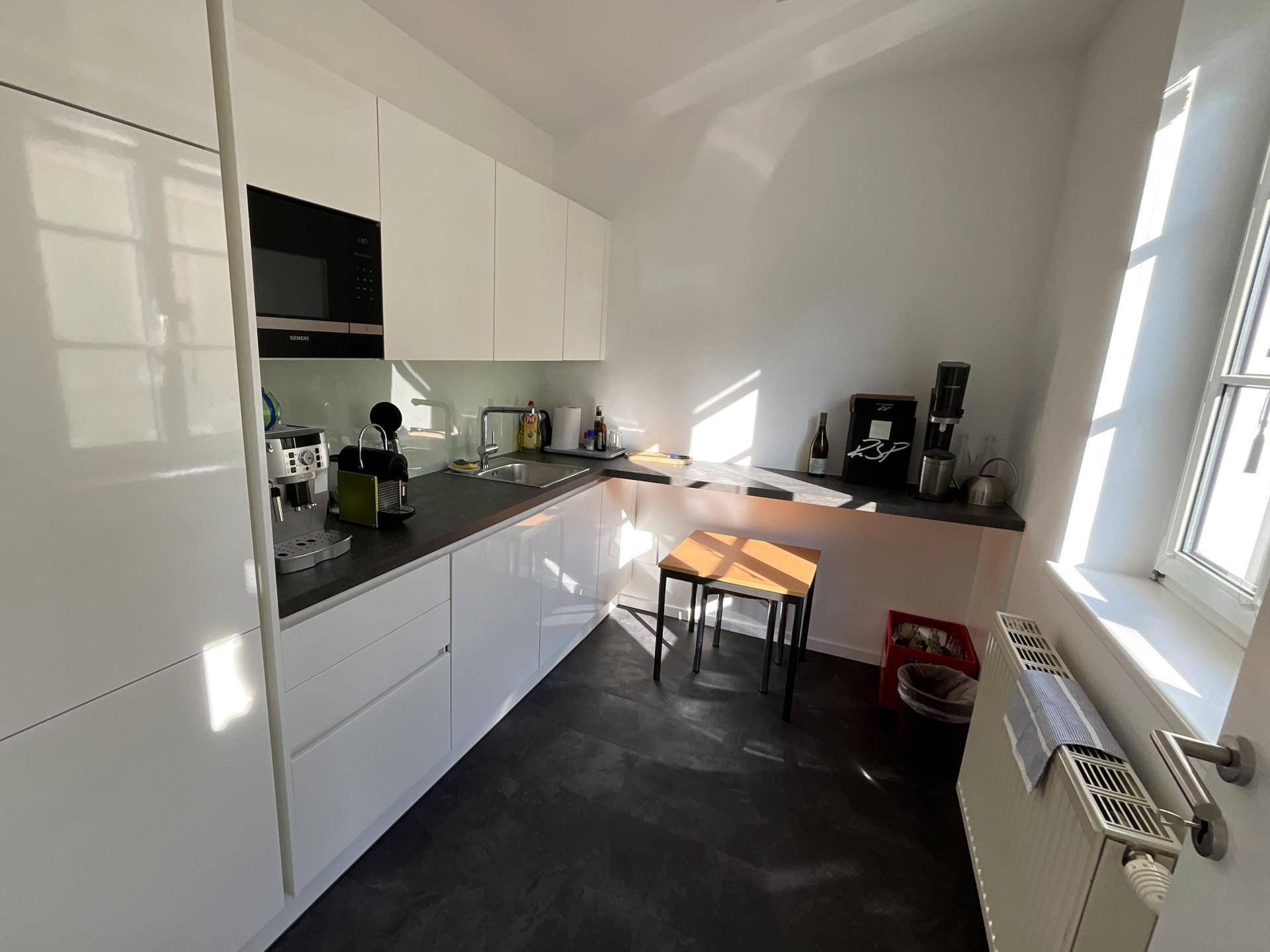 Moderne weiße Küche mit Hochglanzfronten, dunkler Arbeitsplatte und einem Fenster mit viel Tageslicht.