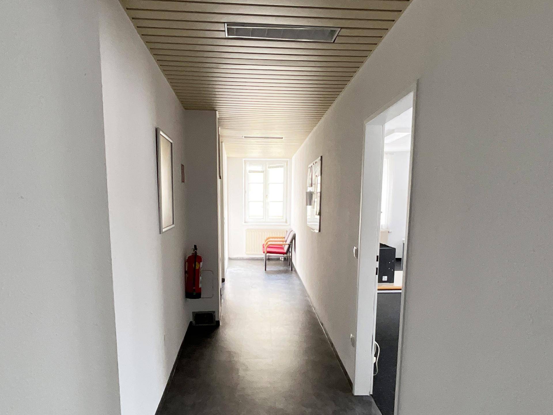 Langer Flur mit Holzdeckenverkleidung, hellem Anstrich und einem Fenster am Ende.