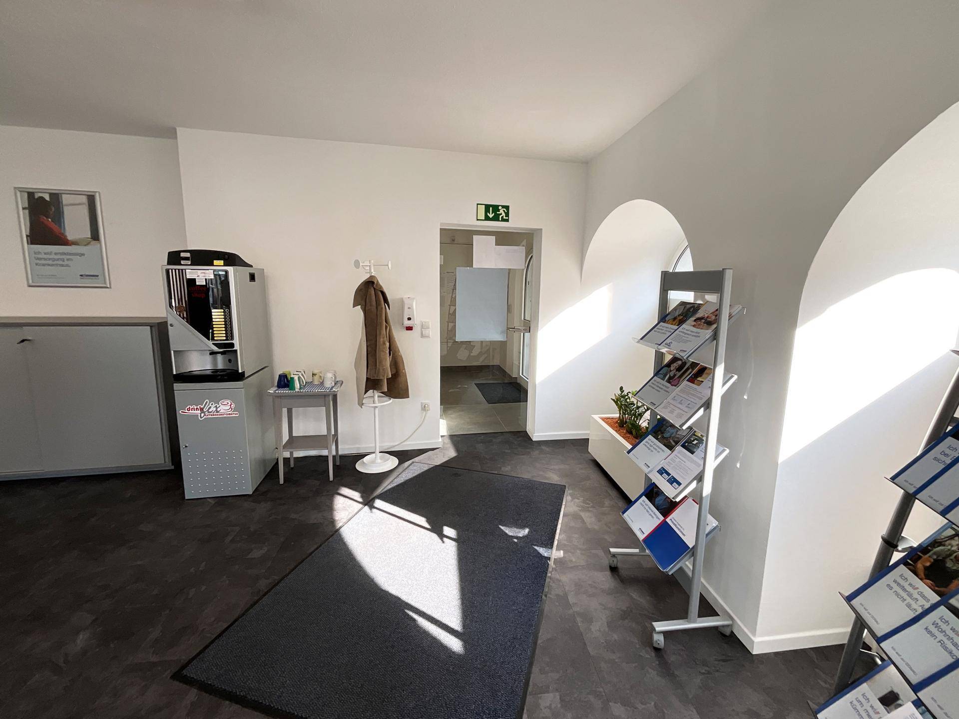 Heller Empfangsbereich mit Kaffeemaschine, Informationsständern und Zugang zu weiteren Büroräumen.