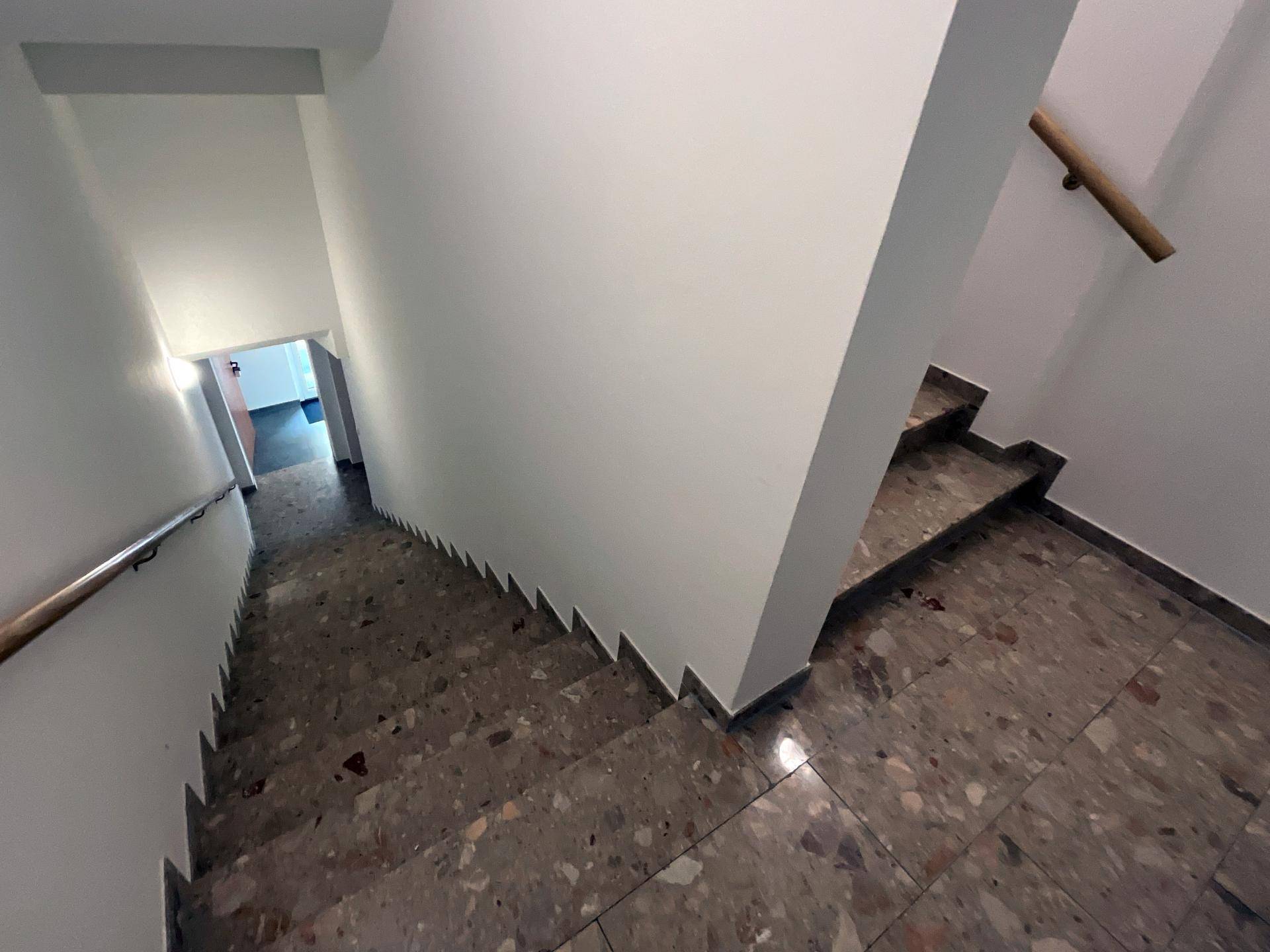 Treppenhaus mit Terrazzoboden und Holzgeländer, das zu den unteren Etagen führt.