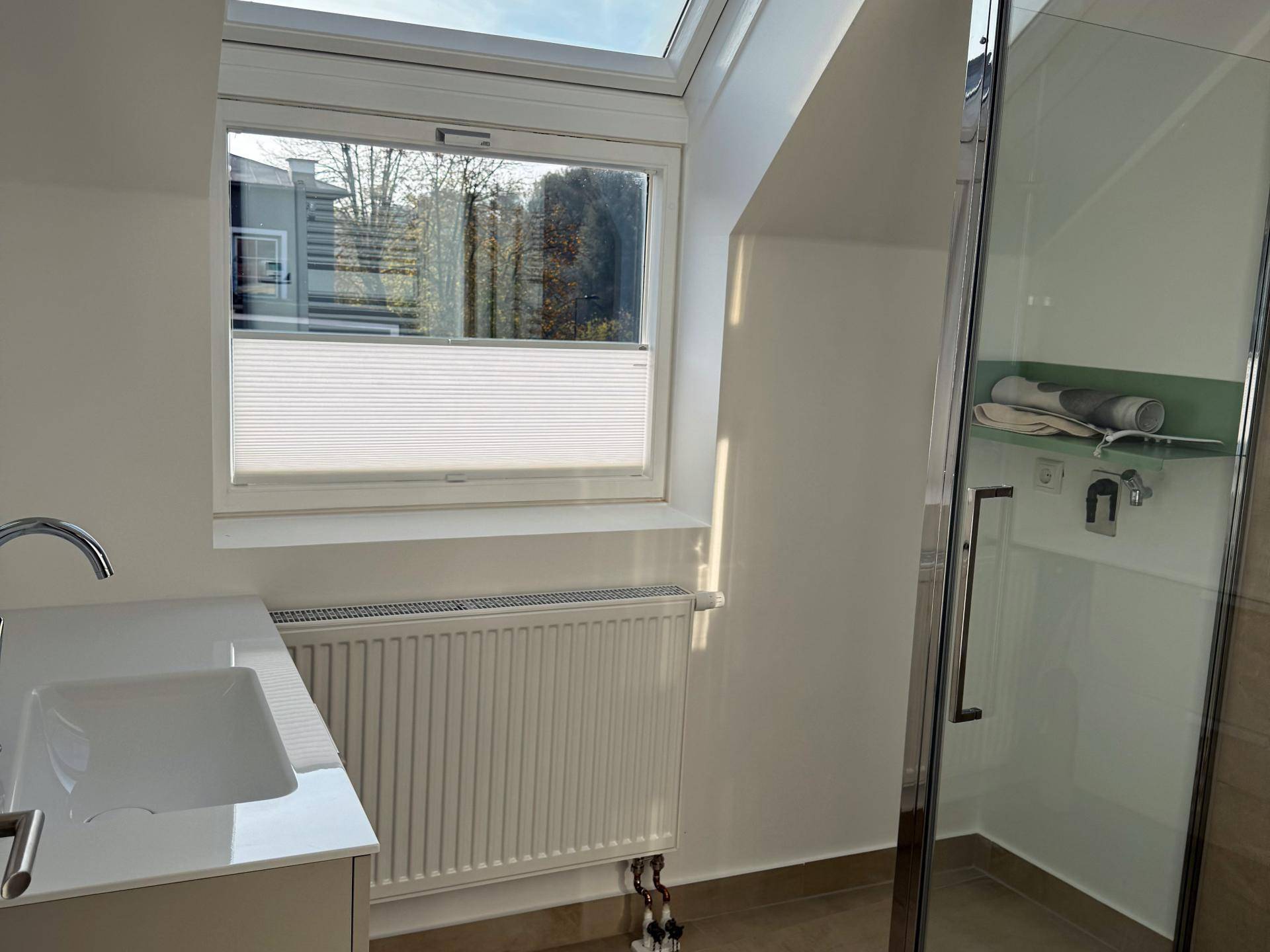 Helles Badezimmer mit Duschkabine, Waschbecken, Heizkörper und einem Fenster, inklusive Dachfenster.