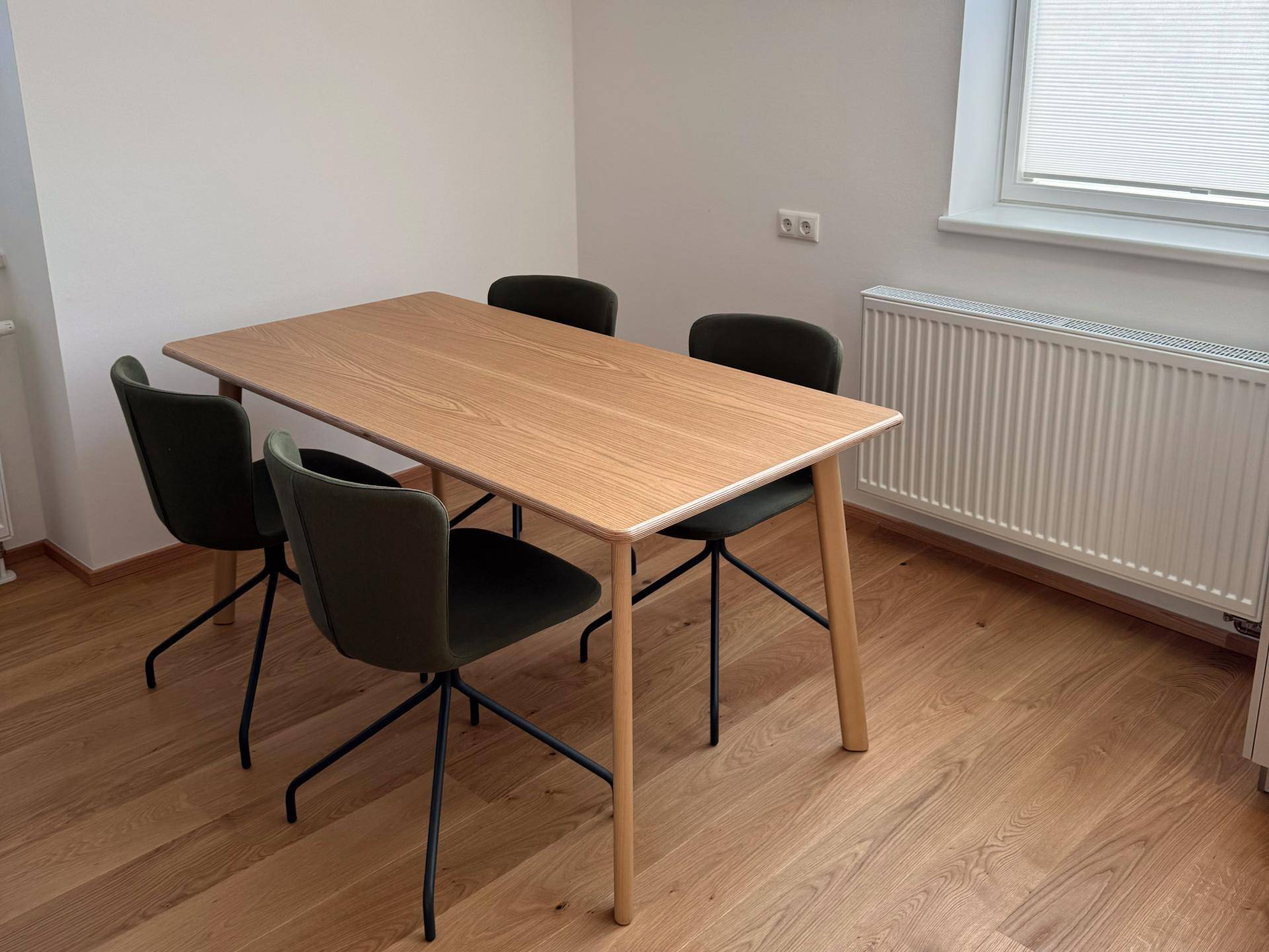 Heller Essbereich mit Holztisch und vier Stühlen, Holzboden und einem Fenster mit Jalousien.