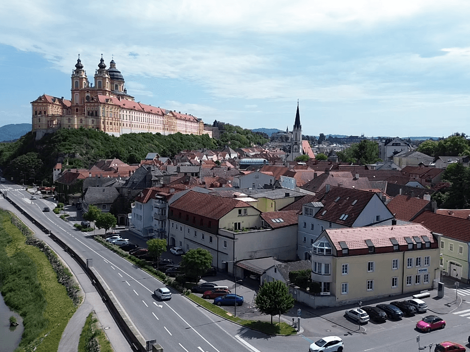 Panoramablick auf eine historische Stadt mit einem imposanten Kloster auf einem Hügel, einem Fluss und grünen Landschaften.
