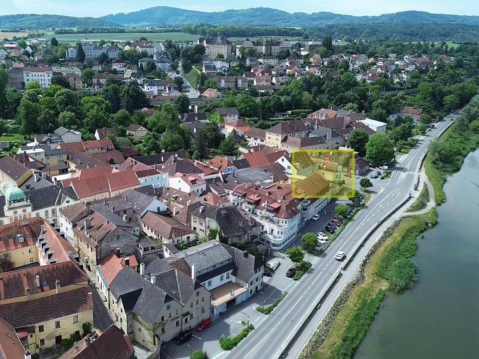 Luftaufnahme einer Stadt am Fluss mit einem hervorgehobenen Gebäude, umgeben von grüner Landschaft und Bergen.