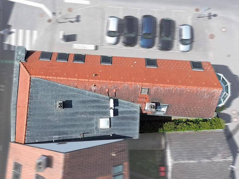 Luftaufnahme eines Gebäudes mit rotem Ziegeldach, Flachdach und mehreren Dachfenstern, Parkplätze sichtbar.