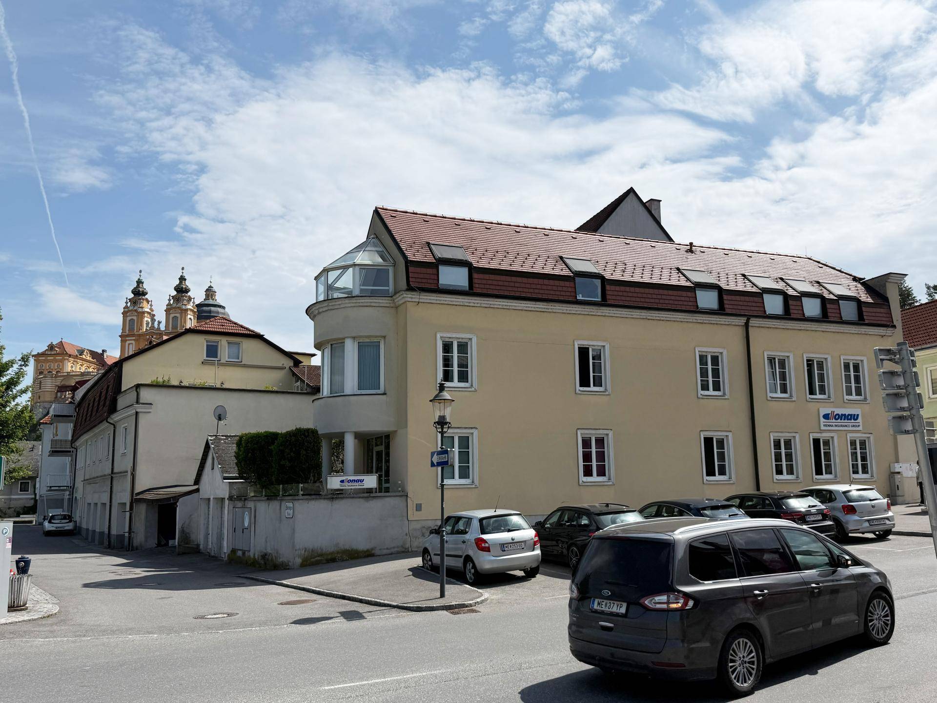 Gelbes Gebäude mit rotem Dach, runder Glaserker und geparkten Autos auf der Straße in einer Stadtansicht.