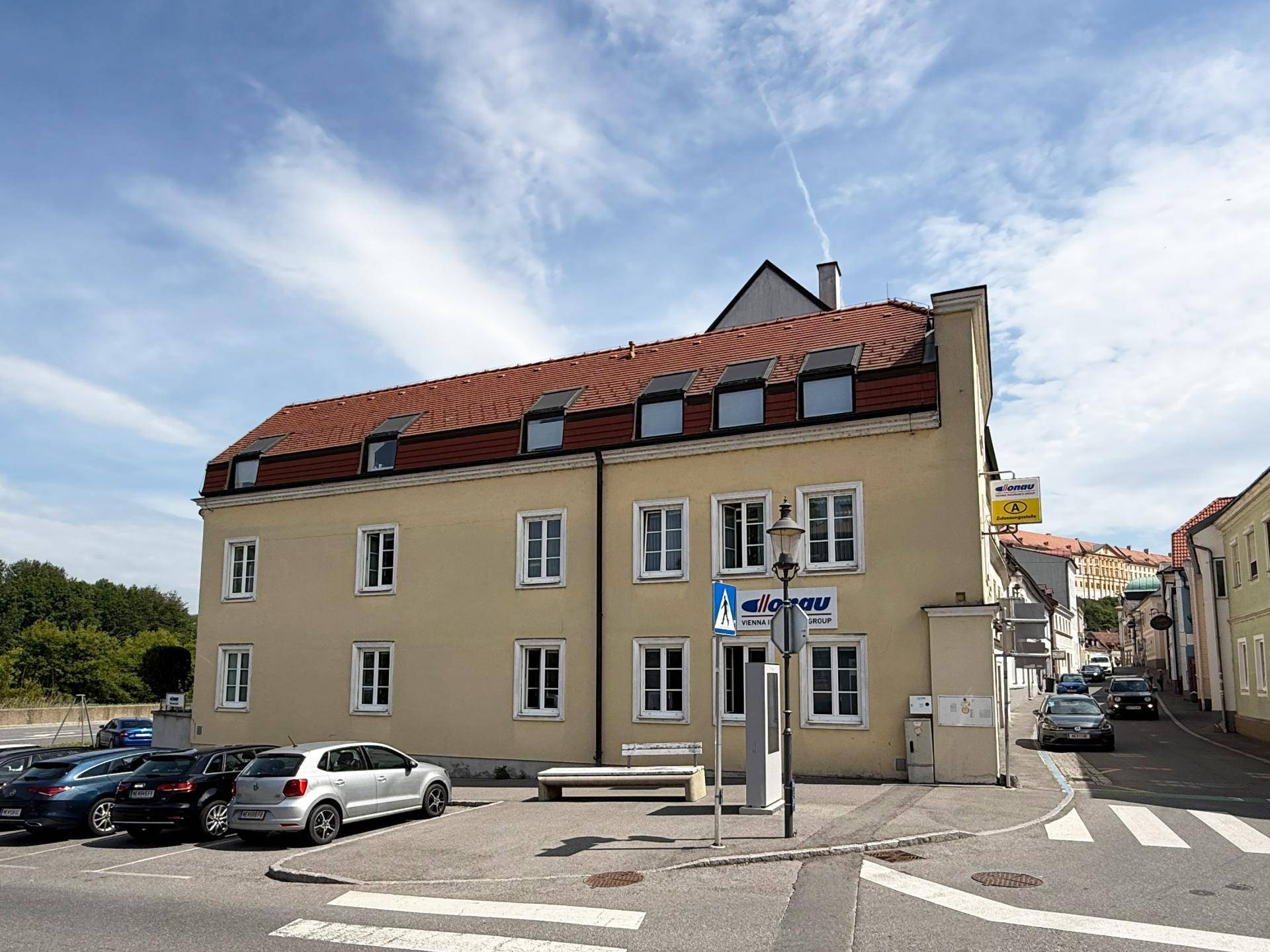 Ansicht des gelben Gebäudes mit rotem Dach und vielen Fenstern, umgeben von geparkten Autos und städtischer Umgebung.