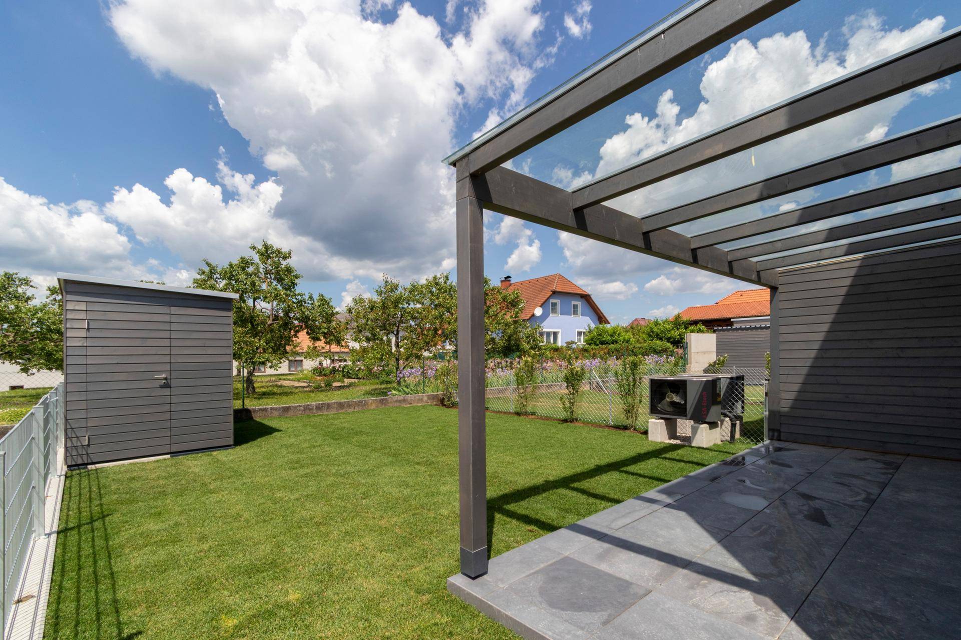 Sonniger Garten mit Rasen, einer gepflasterten Terrasse und einem modernen Gartenhaus.