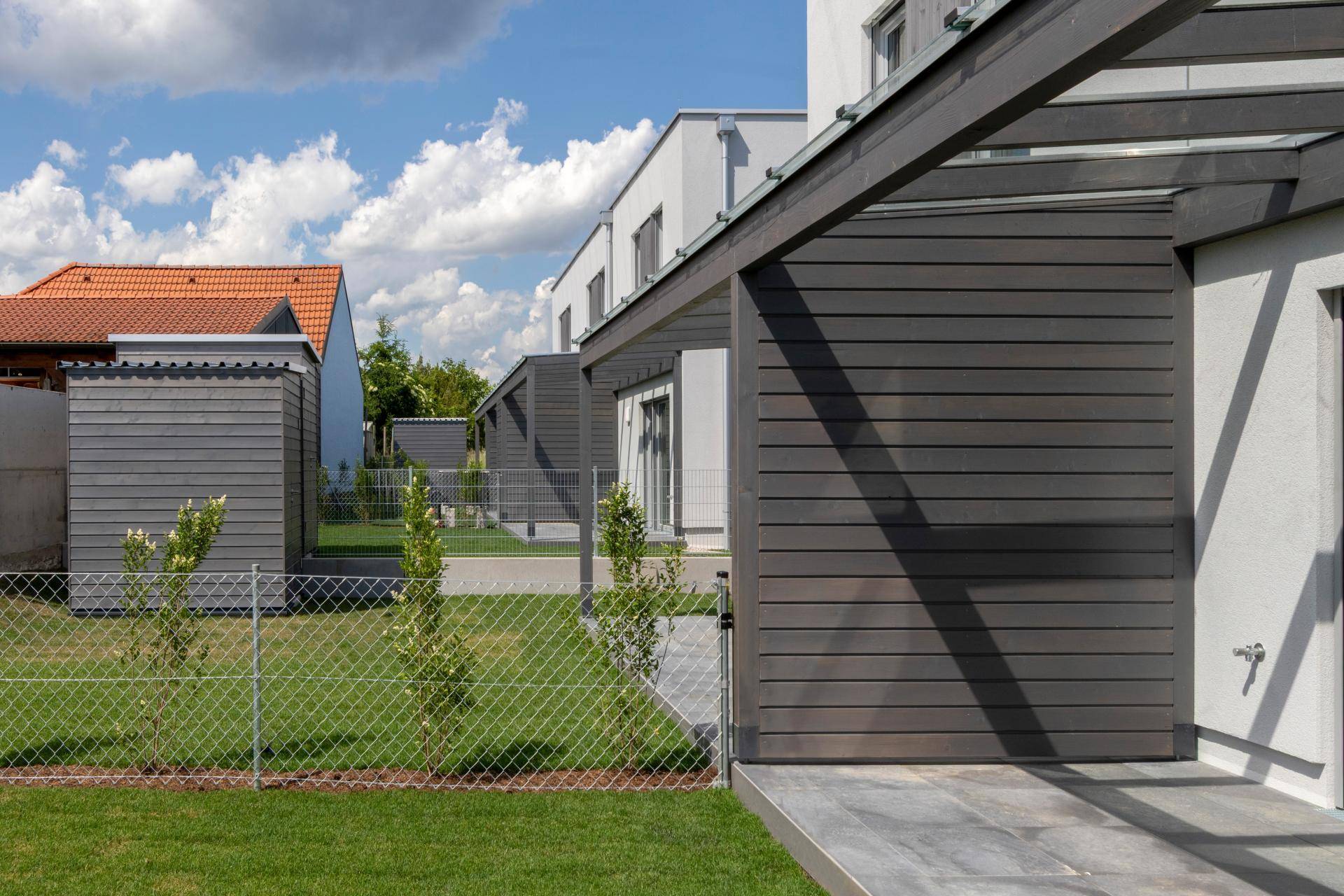Eingezäunter Gartenbereich mit jungen Bäumen und der Seitenansicht eines modernen Reihenhauses.