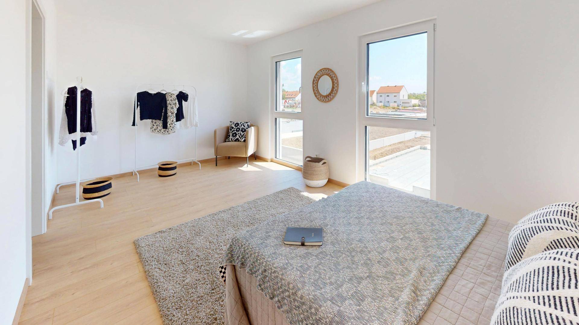 Helles Schlafzimmer mit Doppelbett, Kleiderstange und zwei Fenstern mit Blick auf die Umgebung.