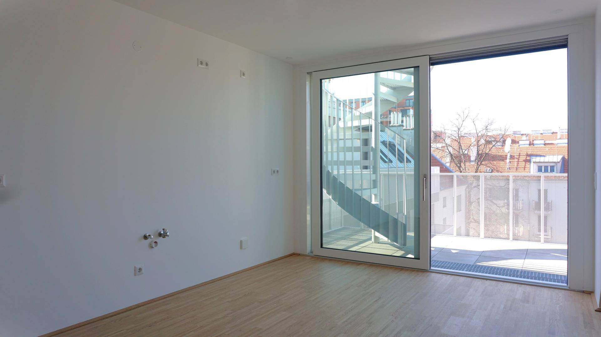Leerer Raum mit Holzboden und großen Fenstern, die Zugang zu einem Balkon mit Wendeltreppe bieten.