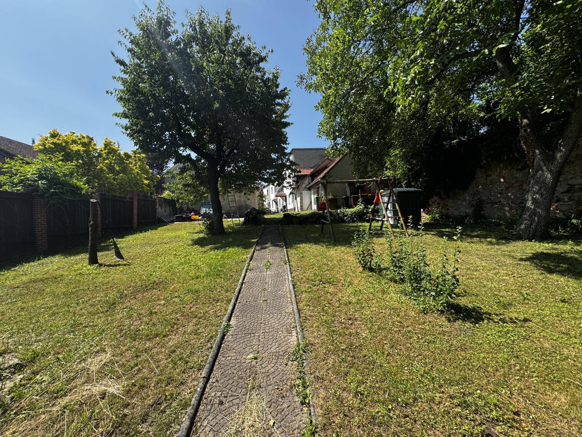 Langer Gartenweg durch eine grüne Rasenfläche, umgeben von Bäumen und einer alten Mauer.