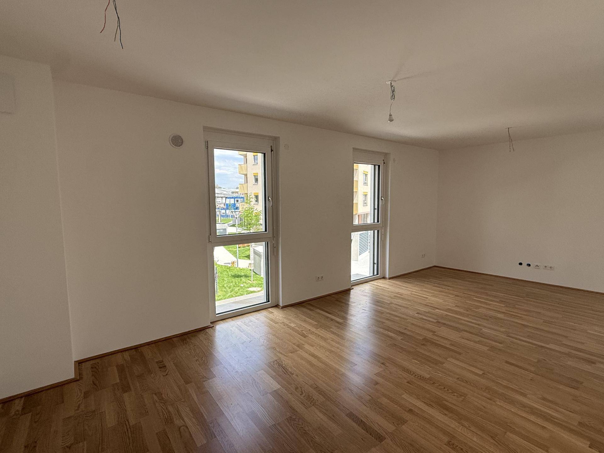 Geräumiger, unmöblierter Wohnraum mit hellem Parkettboden und großen Fenstern mit Blick ins Grüne.