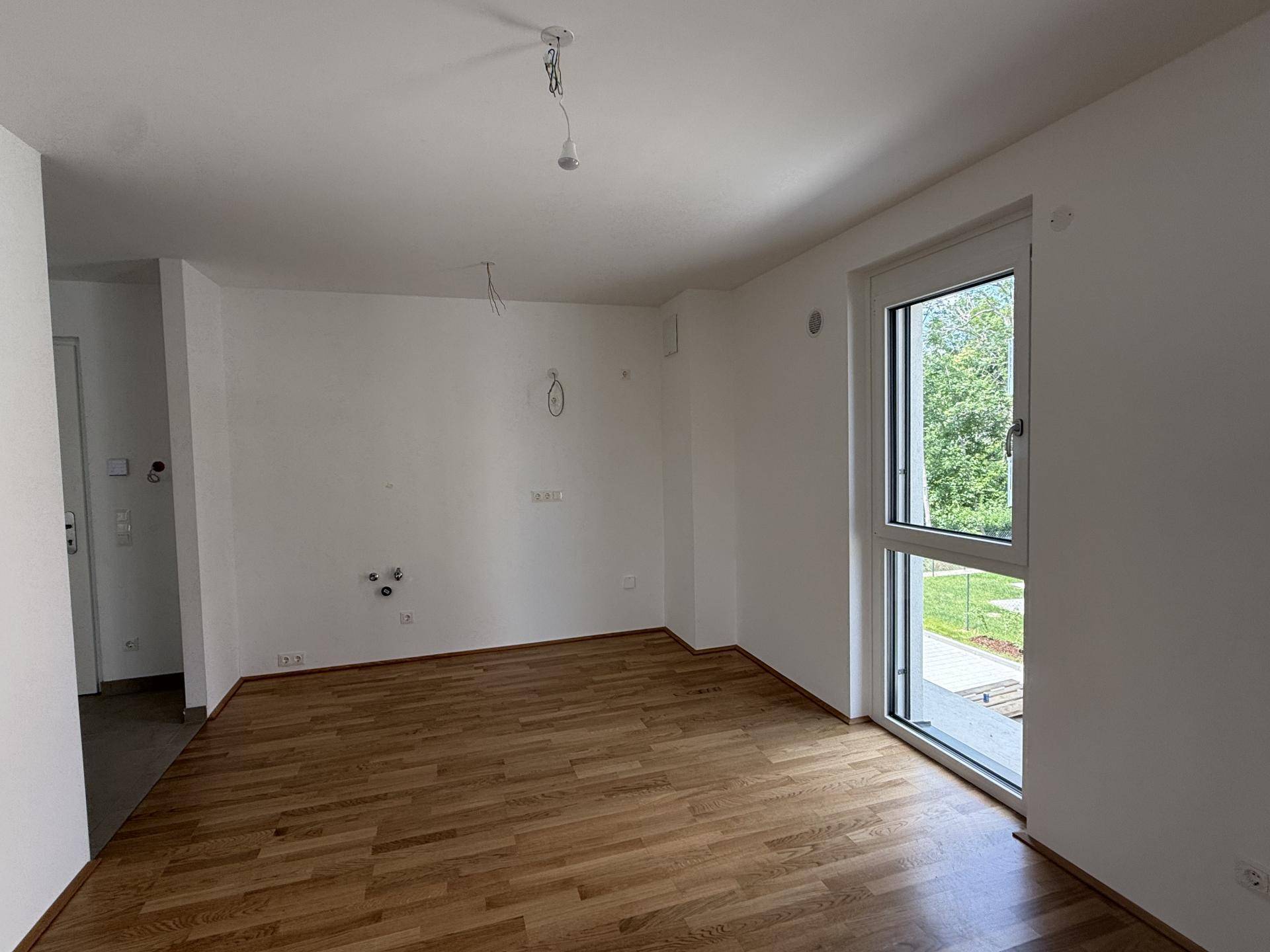 Leerer Küchenbereich mit Parkettboden und Anschlussmöglichkeiten für eine Einbauküche, hell durch großes Fenster.