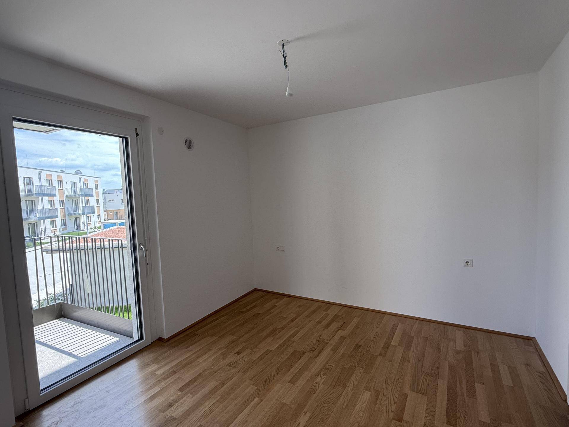 Helles Zimmer mit Parkettboden und Zugang zu einem Balkon, bietet einen Blick auf die umliegenden Gebäude.