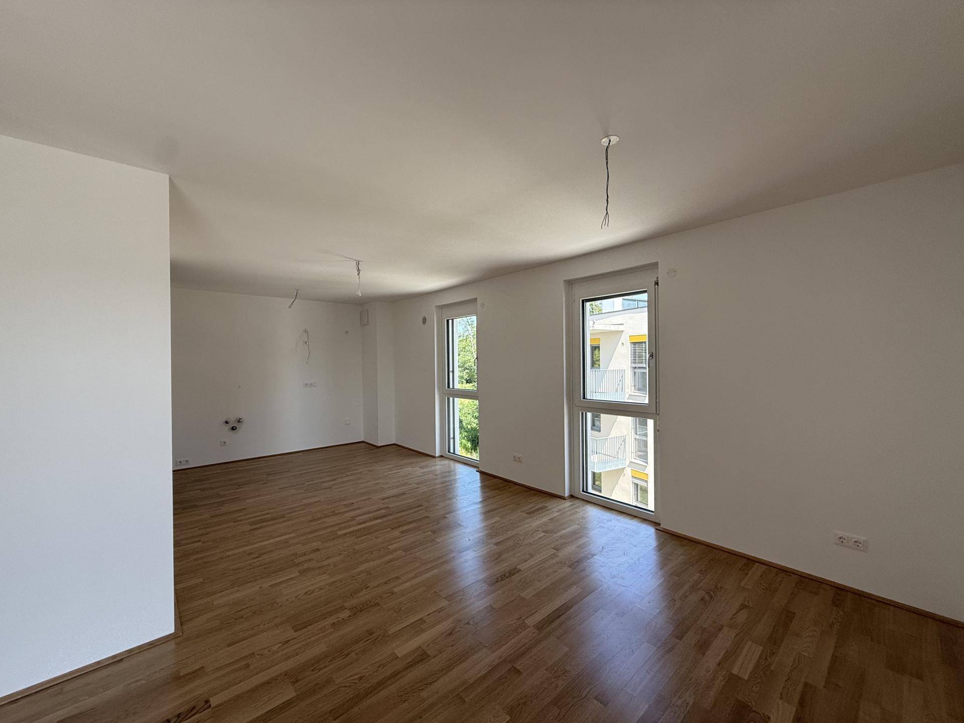 Geräumiges Wohnzimmer mit Holzboden und großen Fenstern, die viel Tageslicht hereinlassen.