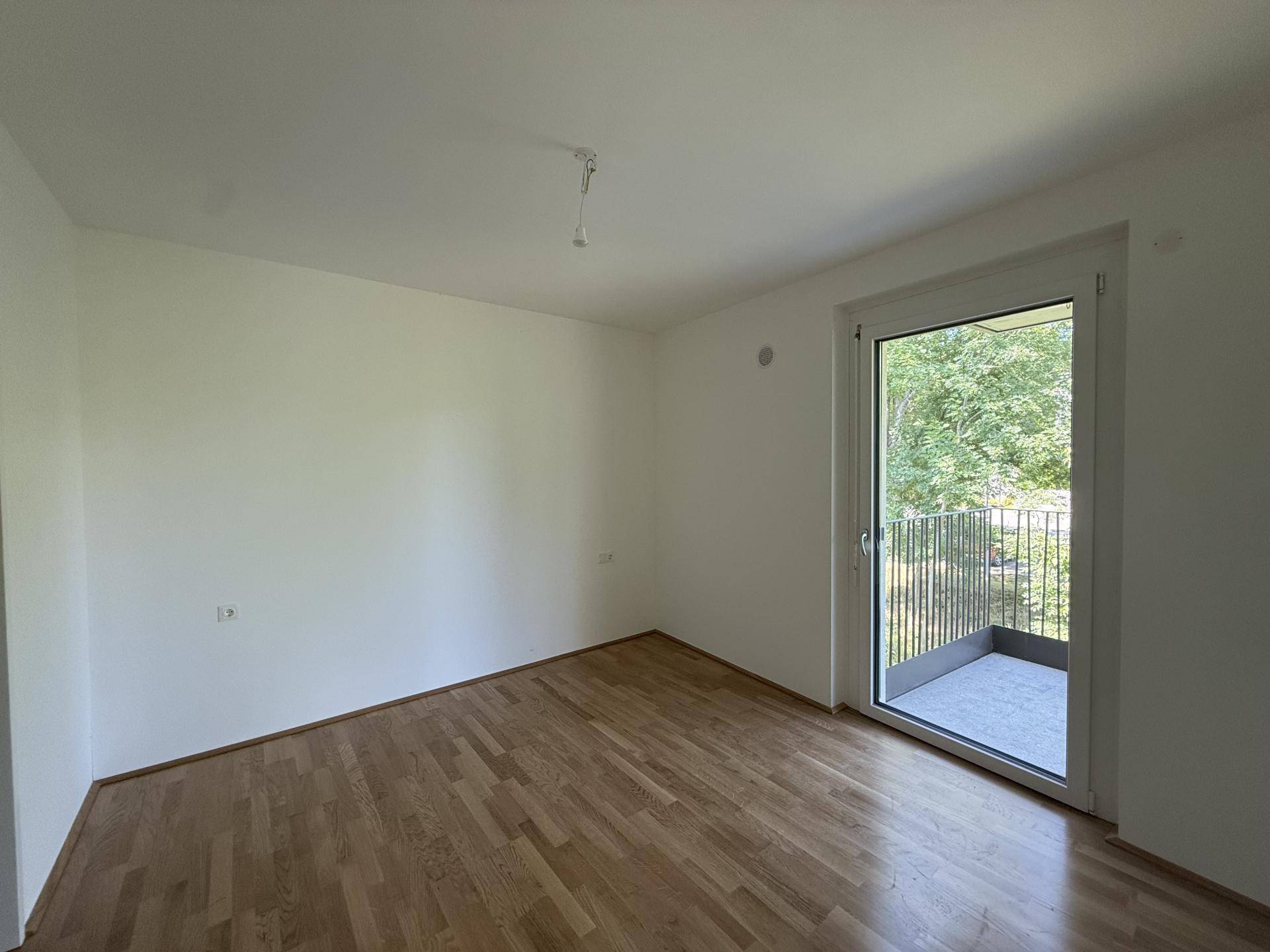 Helles Schlafzimmer mit Parkettboden und Zugang zu einem Balkon mit Blick ins Grüne.